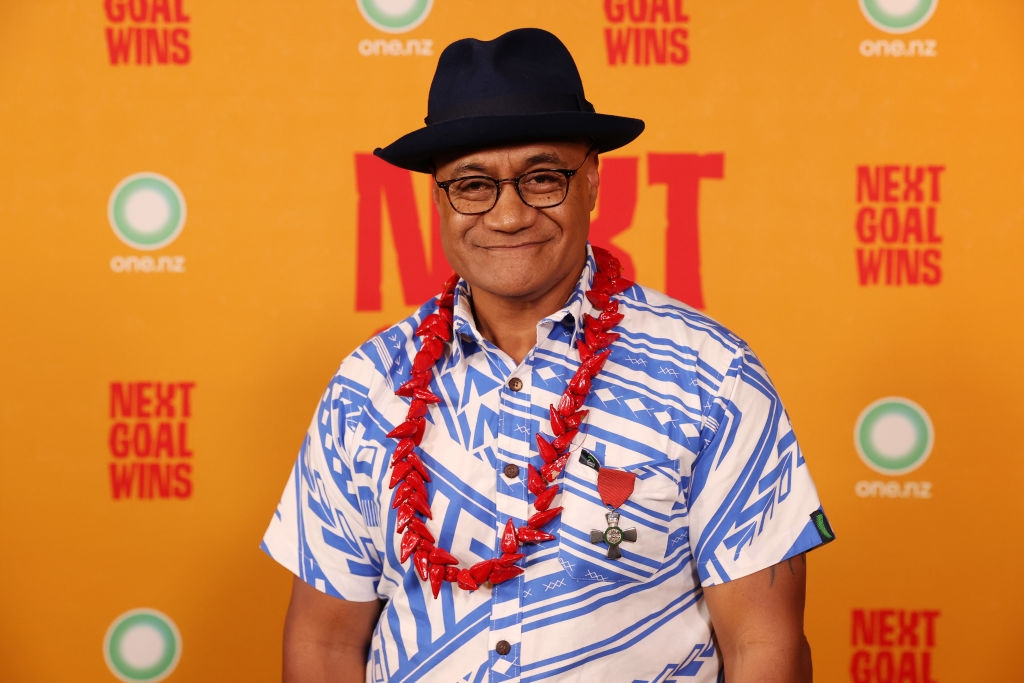 Person wearing a blue and white patterned shirt, a black hat, and a red flower lei, standing in front of a "Next Goal Wins" backdrop