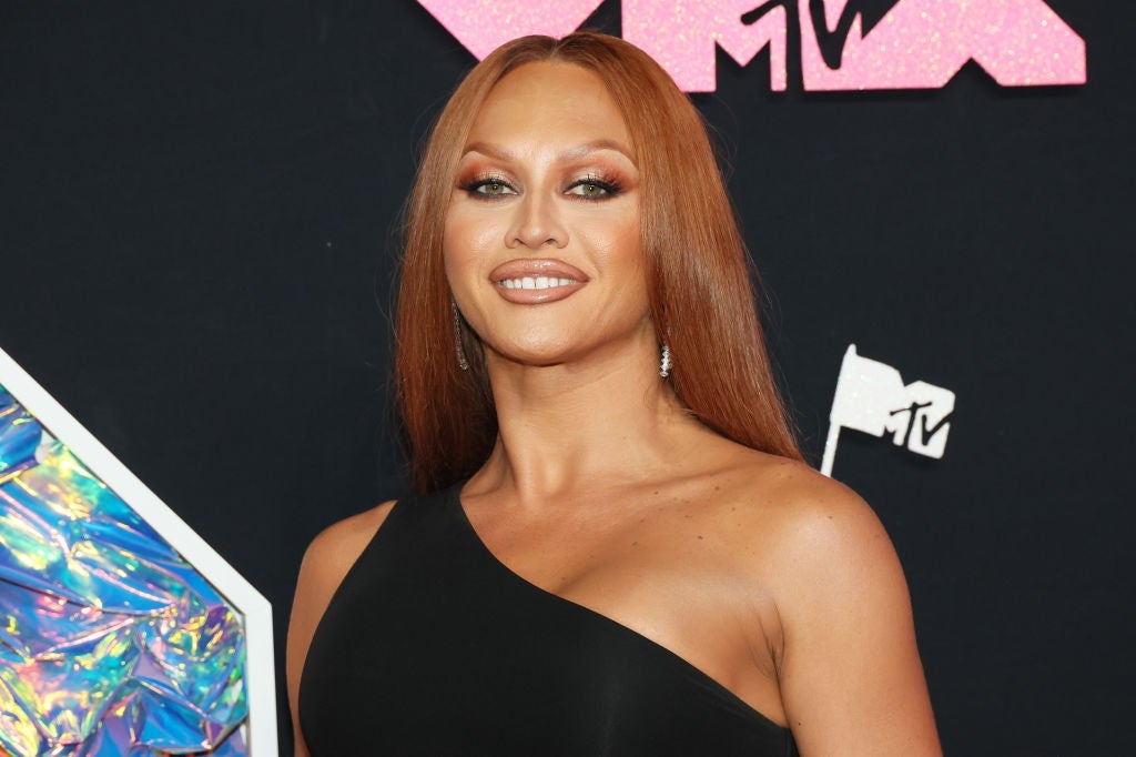 Person with long hair wearing a one-shoulder dress poses confidently at an event. MTV logo visible in the background