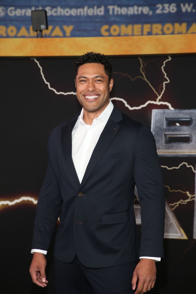 Person in a tailored suit smiling at a theater event. Background shows a partially visible theater sign