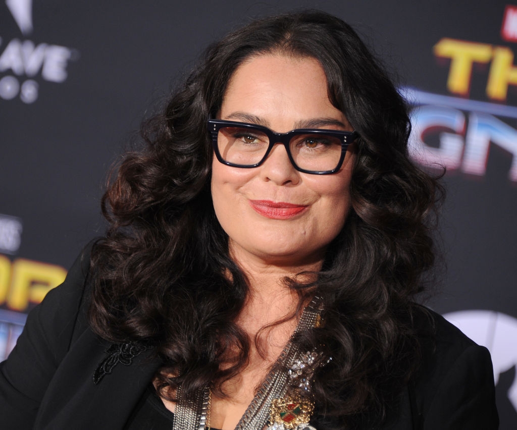 Person with curly hair and glasses on a red carpet, wearing a black outfit with layered necklaces