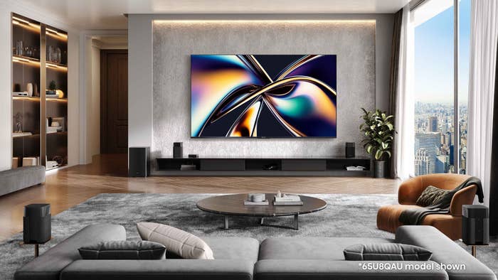 Spacious modern living room with large flat-screen TV displaying abstract art. Cityscape visible through floor-to-ceiling windows.