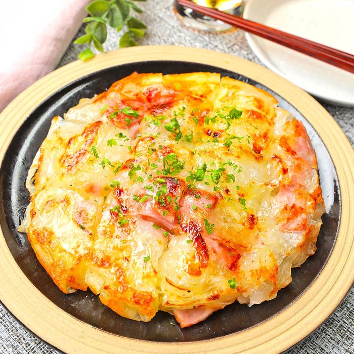 お好み焼きスタイルの玉ねぎ料理が皿に盛られている。上にパセリが散らされ、ハムやチーズが見える。隣に箸と皿。