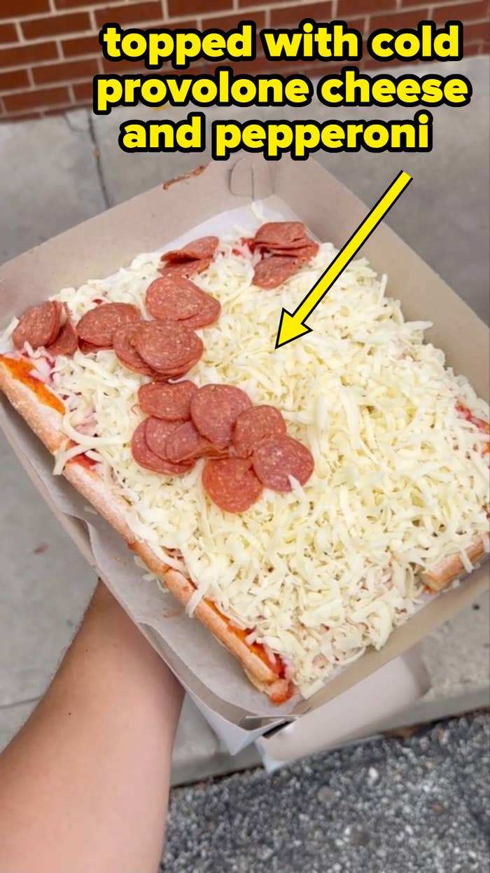 A hand holding a tray with a rectangular pizza topped with shredded cheese and pepperoni slices, against a brick wall and sidewalk background