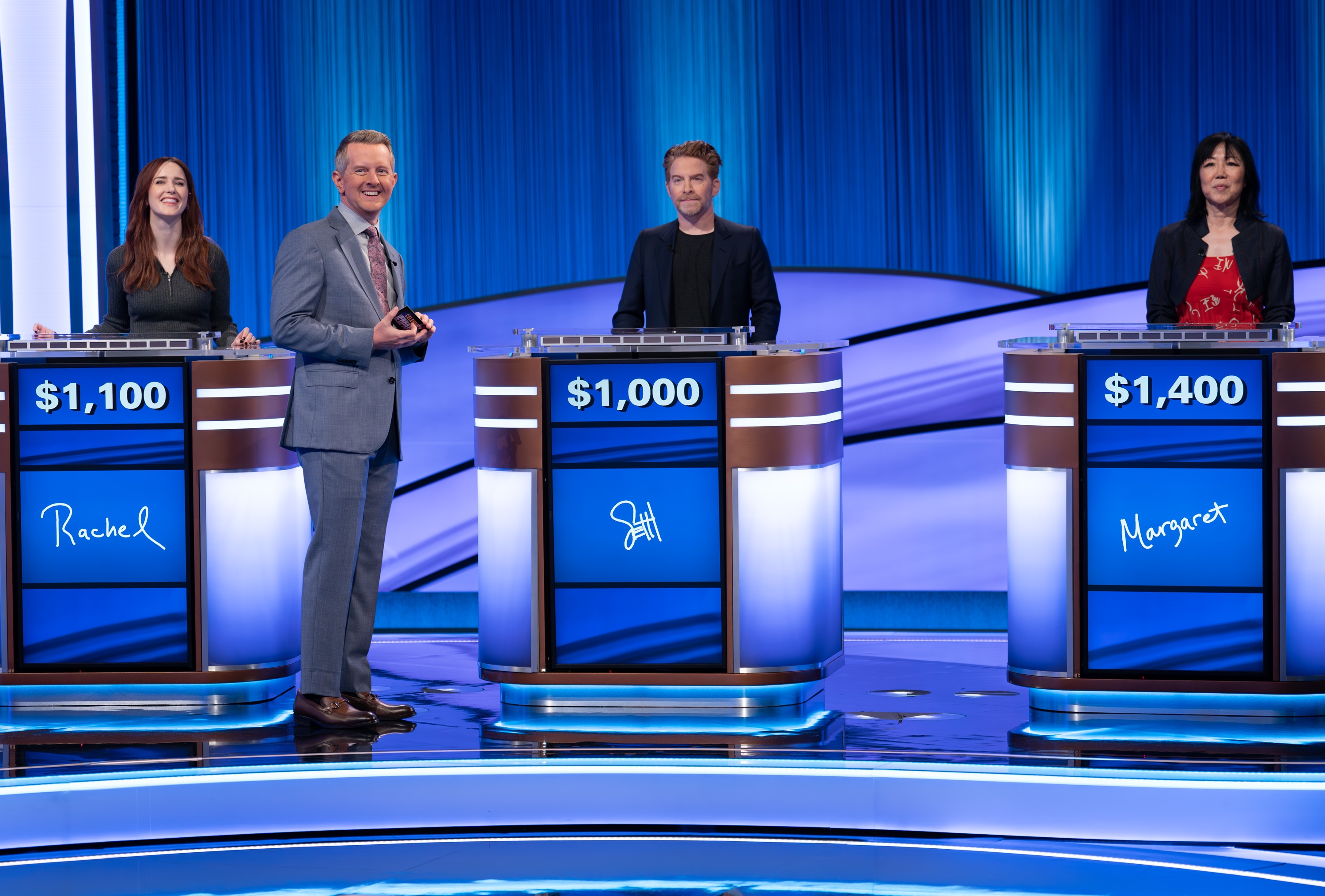 Four game show contestants stand behind podiums displaying their scores and names: Rachel, Jill, and Margaret, with the host in a suit beside them