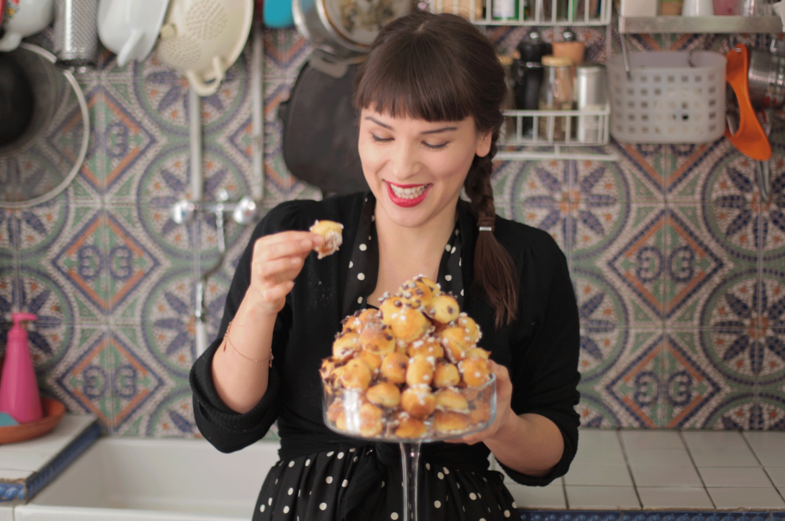 A smiling person in a kitchen holds a plate of pastries, wearing a polka dot dress