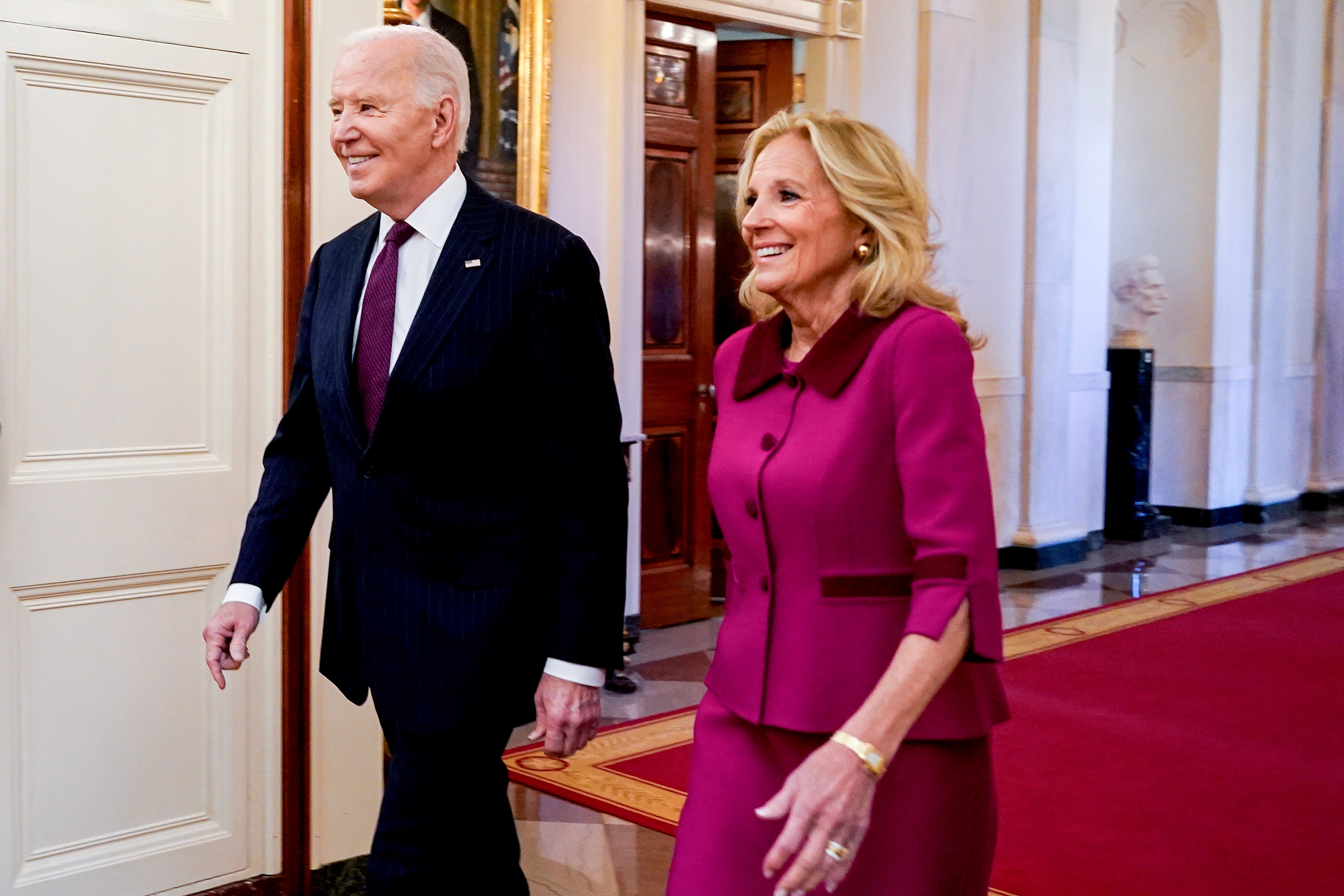 A man in a suit and a woman in a matching skirt suit walk together down a hall with a red carpet