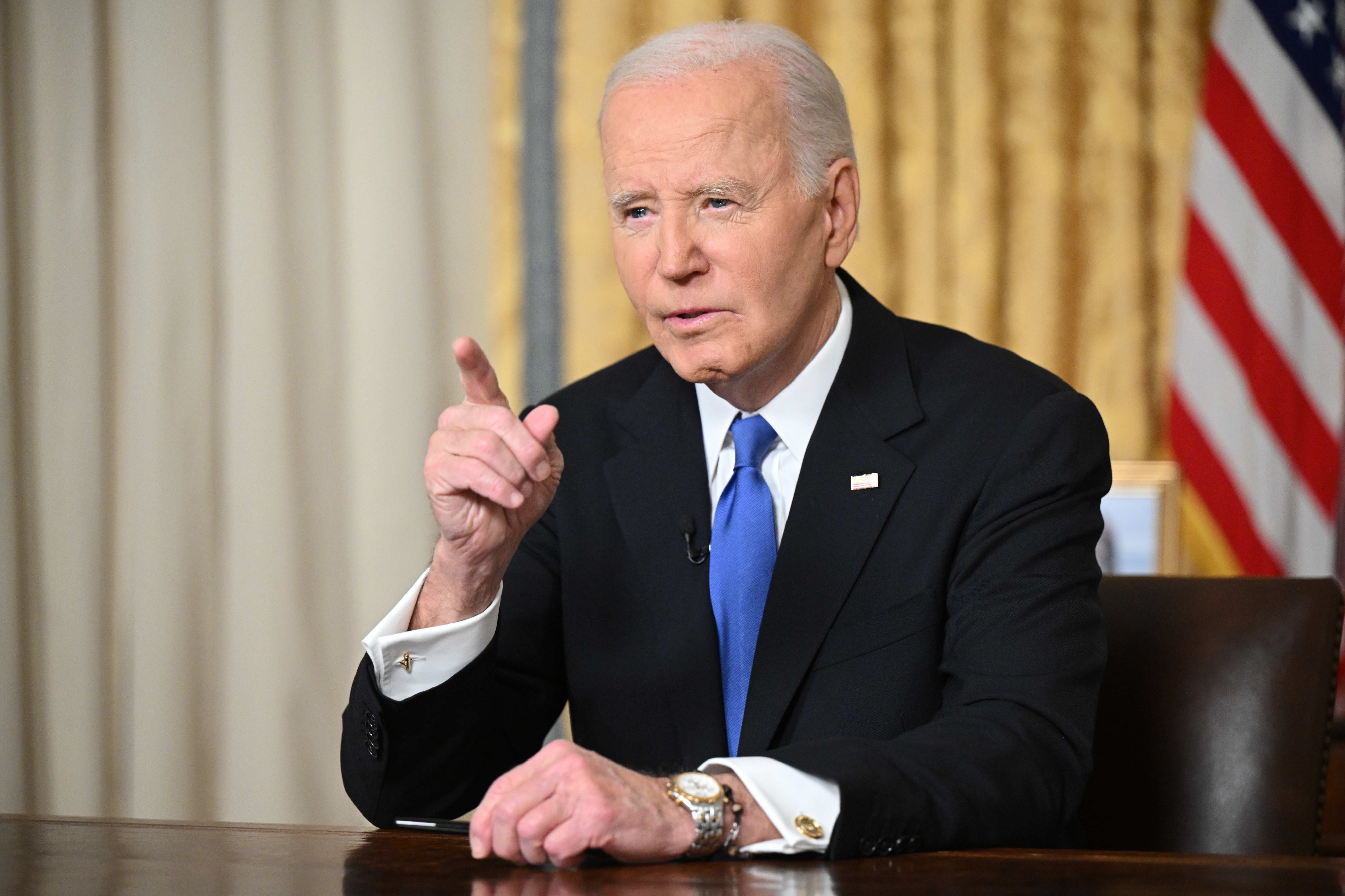 A man in a suit and tie sits at a table, gesturing with his hand, with a flag in the background