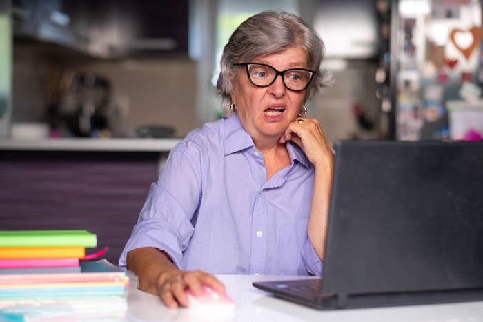 Elderly pistillate   with glasses, amazed  expression, looking astatine  a laptop screen. Books and folders connected  the array  beside her