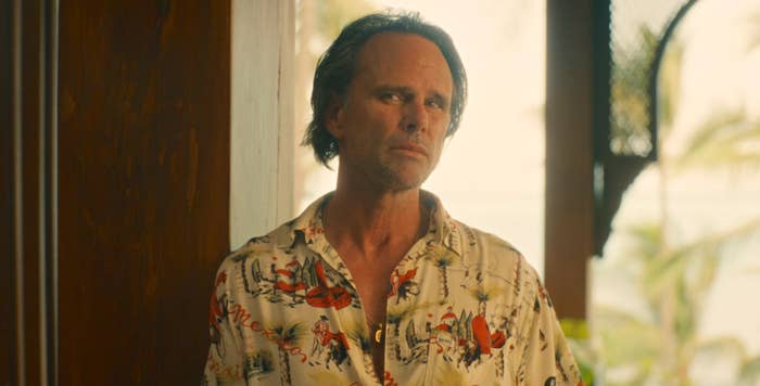 Man in floral shirt leans against a wall, looking thoughtful
