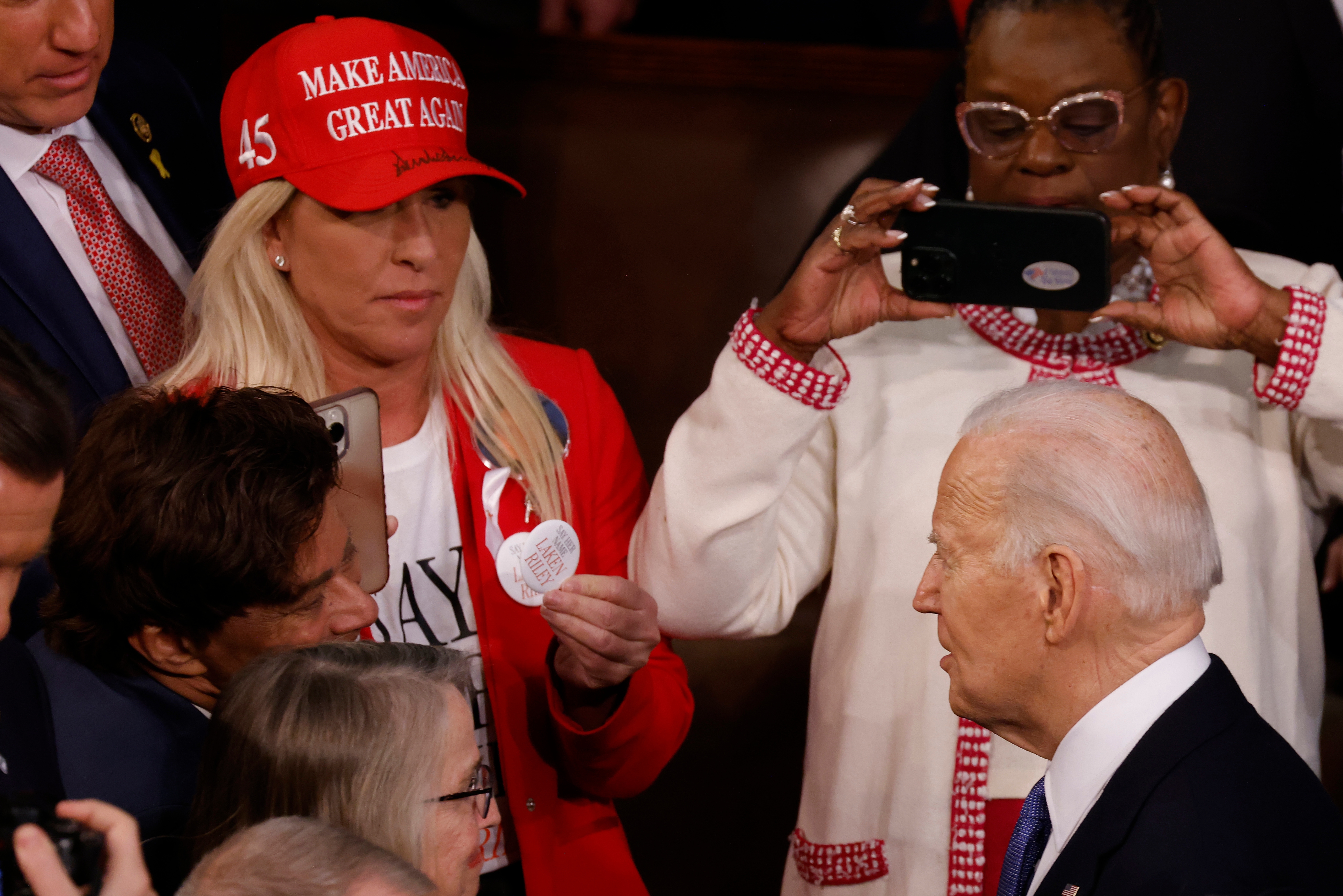 A woman in a "Make America Great Again" cap shows a paper to President Biden at an event, while others around them take photos and watch