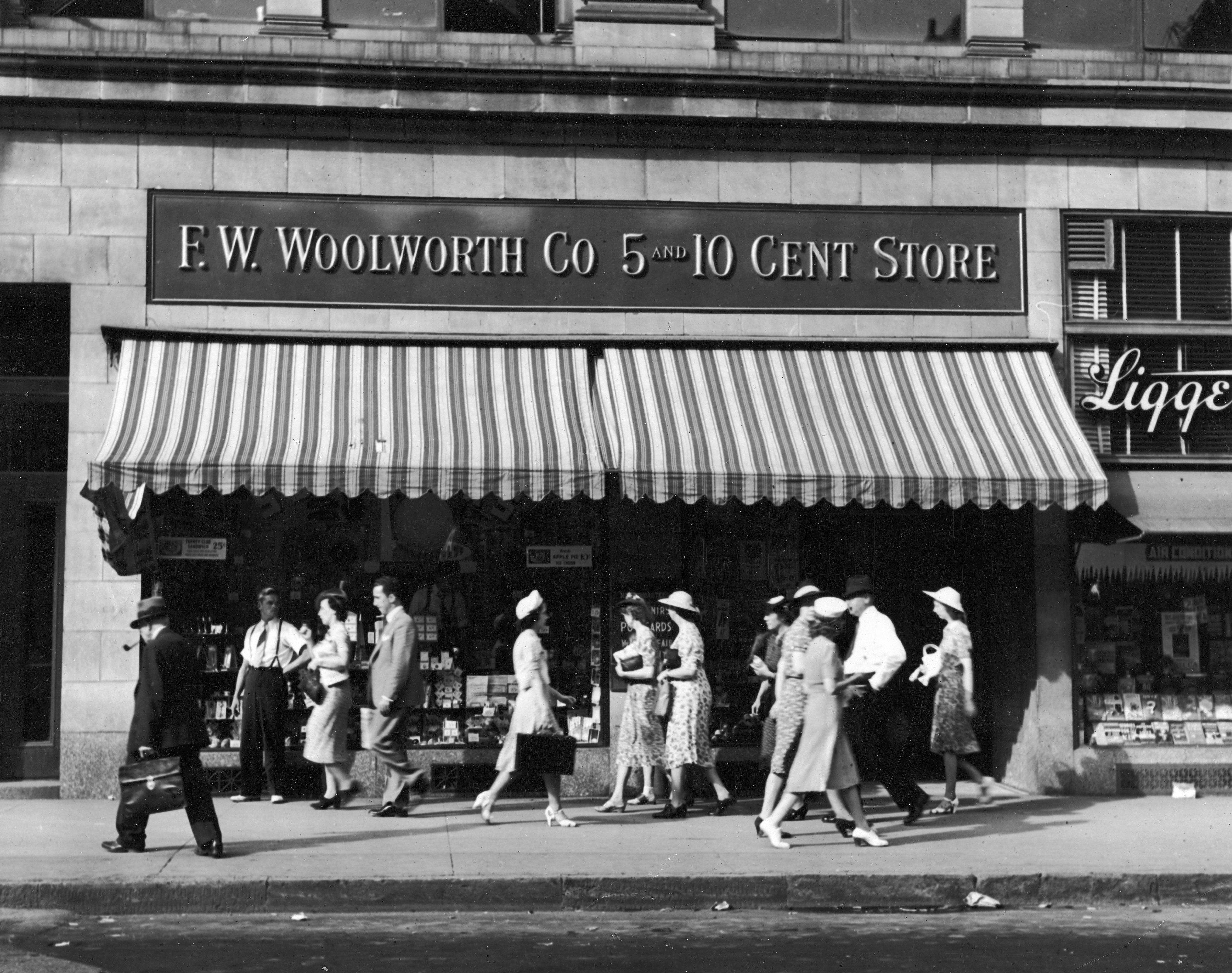 People walk past a F.W. Woolworth Co. 5 and 10 Cent Store, featuring a striped awning, in a bustling street scene from the early 20th century