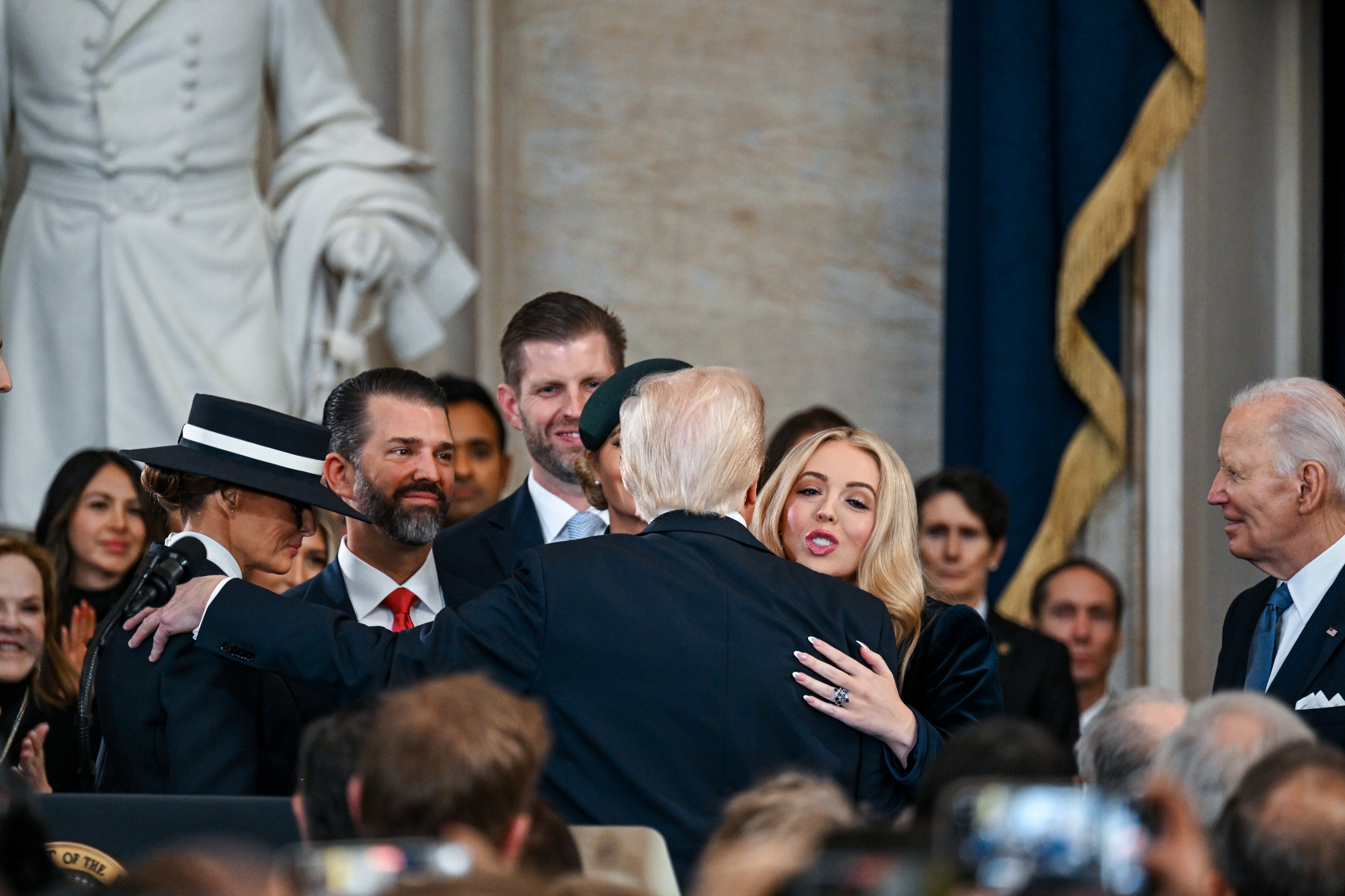 A group of people, including a well-known political figure, in a formal setting. A woman greets the political figure with a hug amidst the gathering