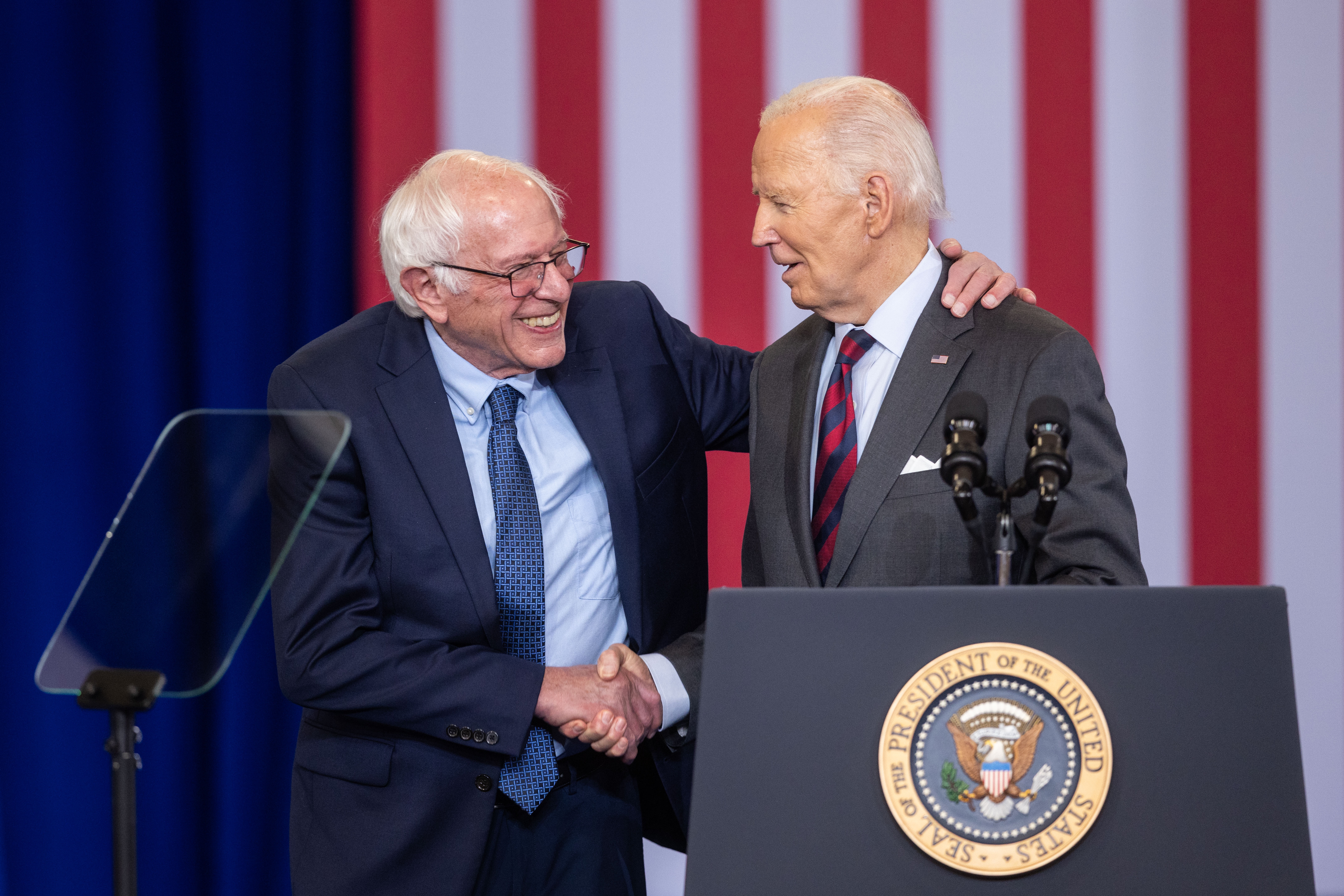 Two senior politicians shake hands and smile at a podium with U.S. emblems. A background with vertical stripes is visible behind them