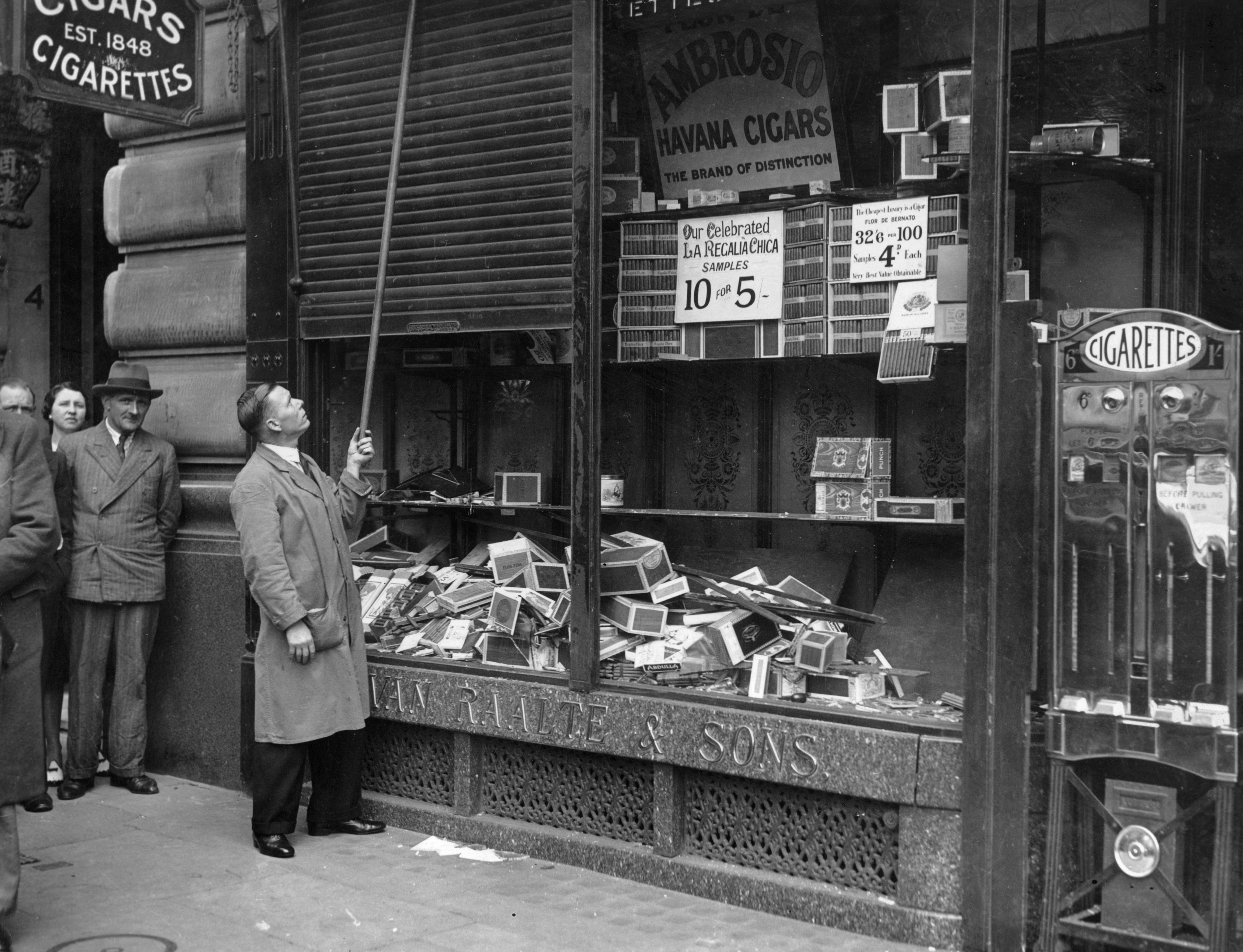 Man using a pole to close a shop shutter with displays of cigar boxes on the street below. Two men observe from the sidewalk
