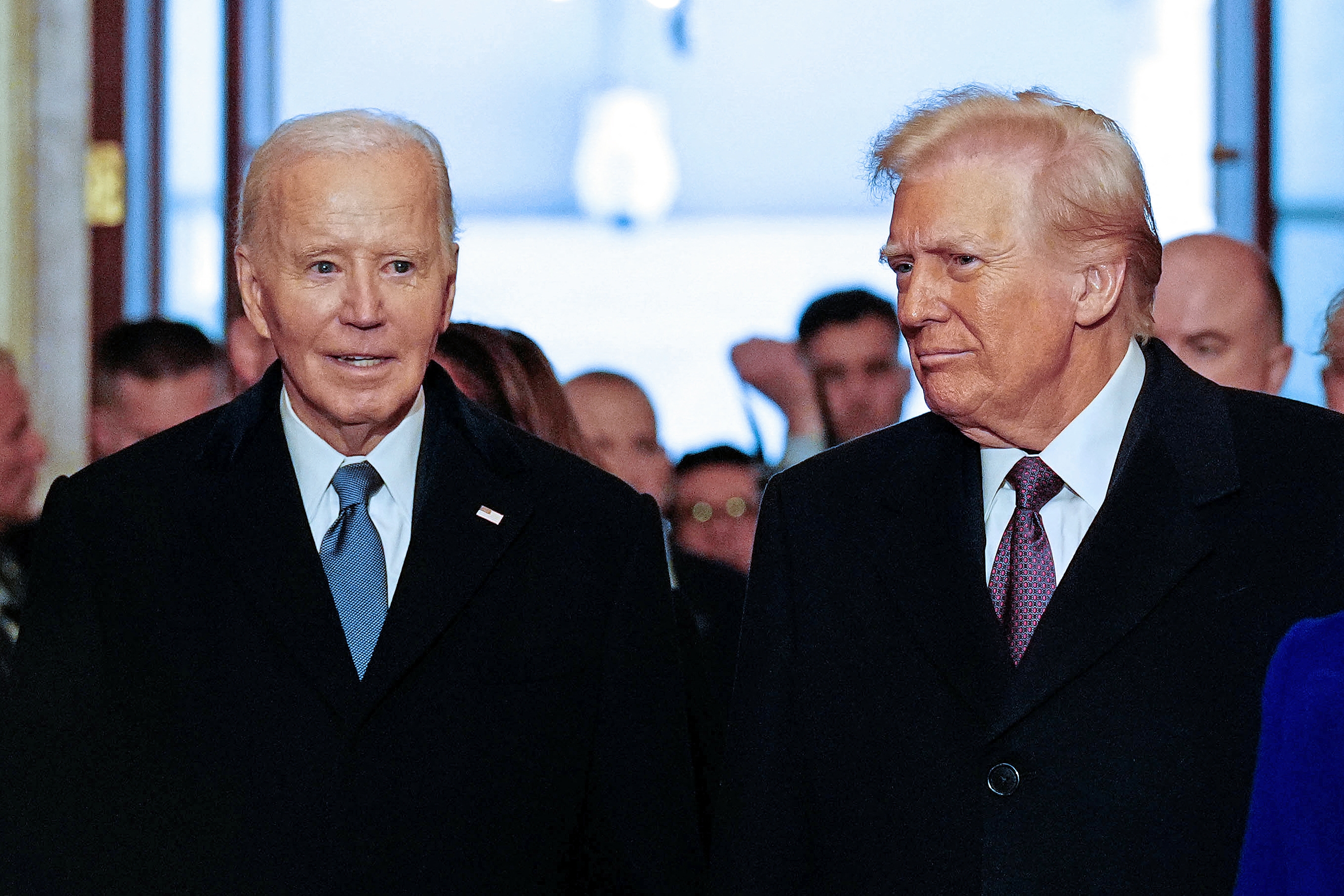 Two men in formal suits stand together at a public event, engaged in conversation. A crowd is visible in the background