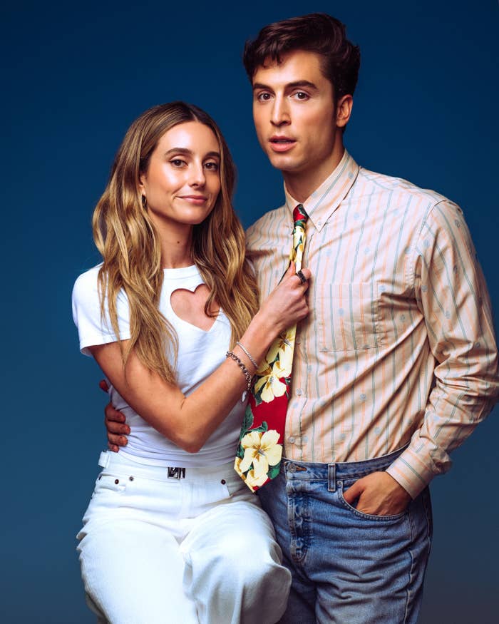 Two people pose together. One wears a heart-cutout top and white jeans, while the other wears a striped shirt and floral tie