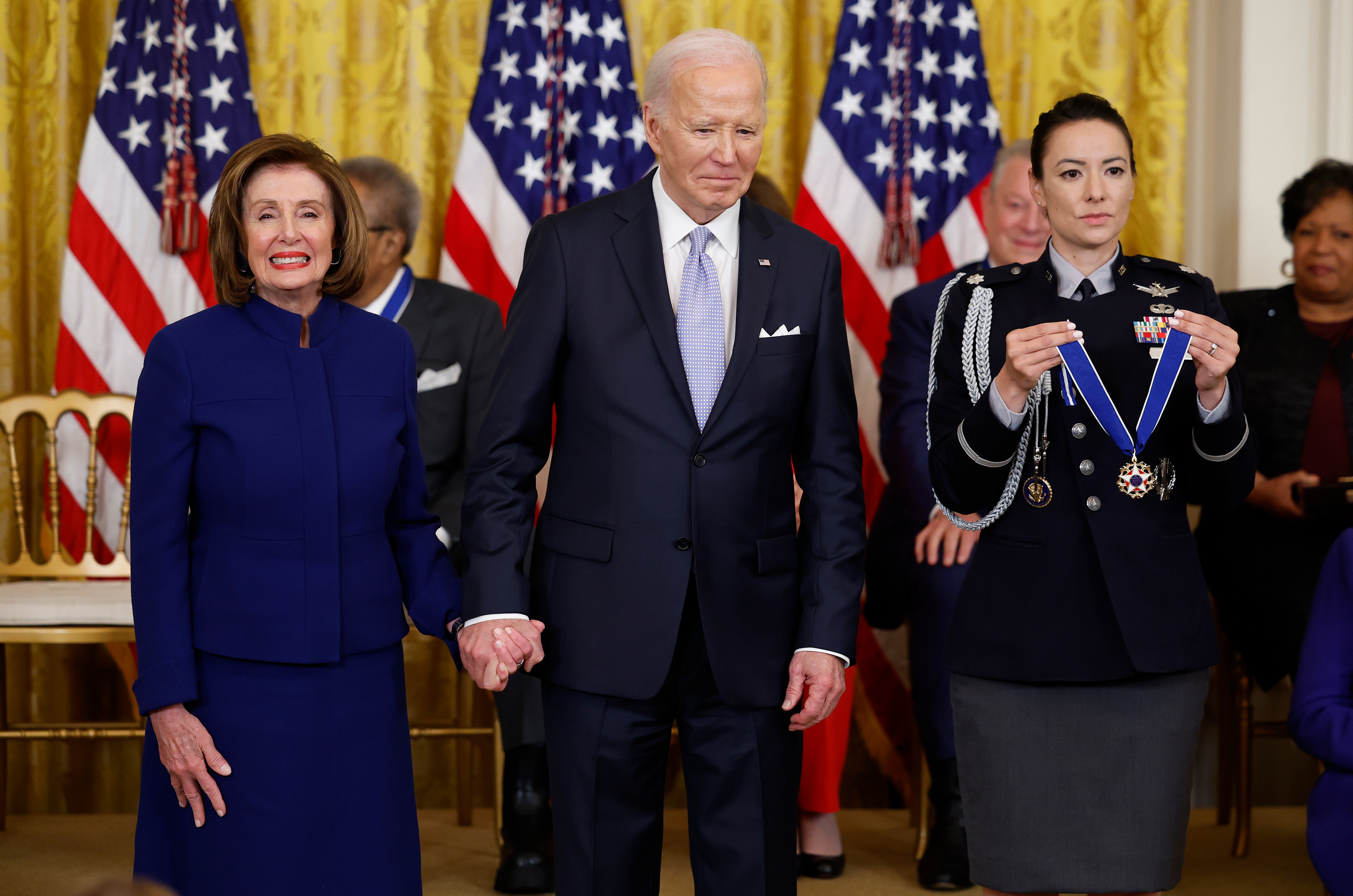 A formal event with a man in a suit holding hands with a woman. A uniformed officer is presenting a medal. Several flags are displayed in the background