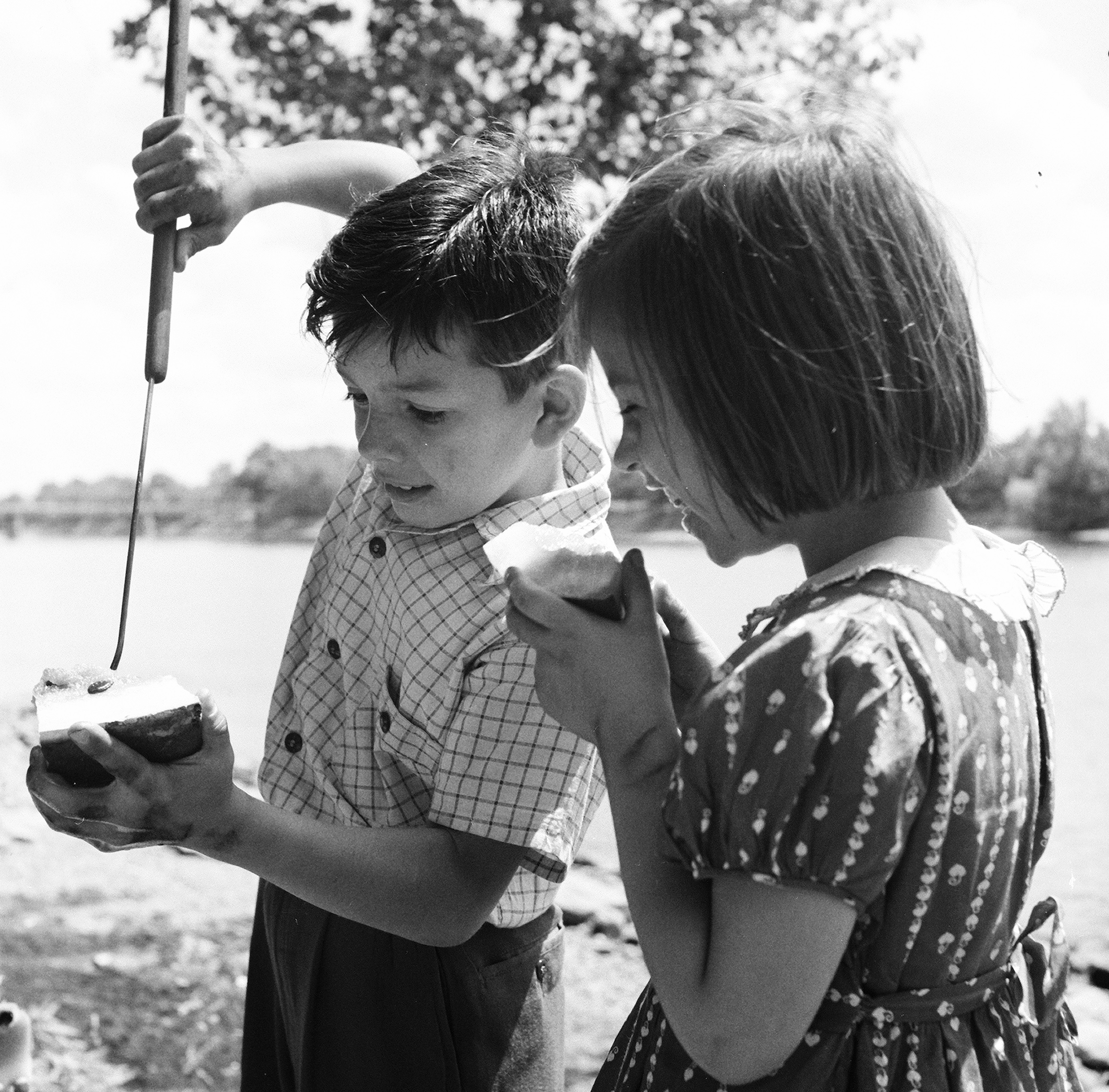 Two children enjoying a summer day by a lake, one pours a drink into a cup, and the other holds a cup, both smiling and engaged in the moment