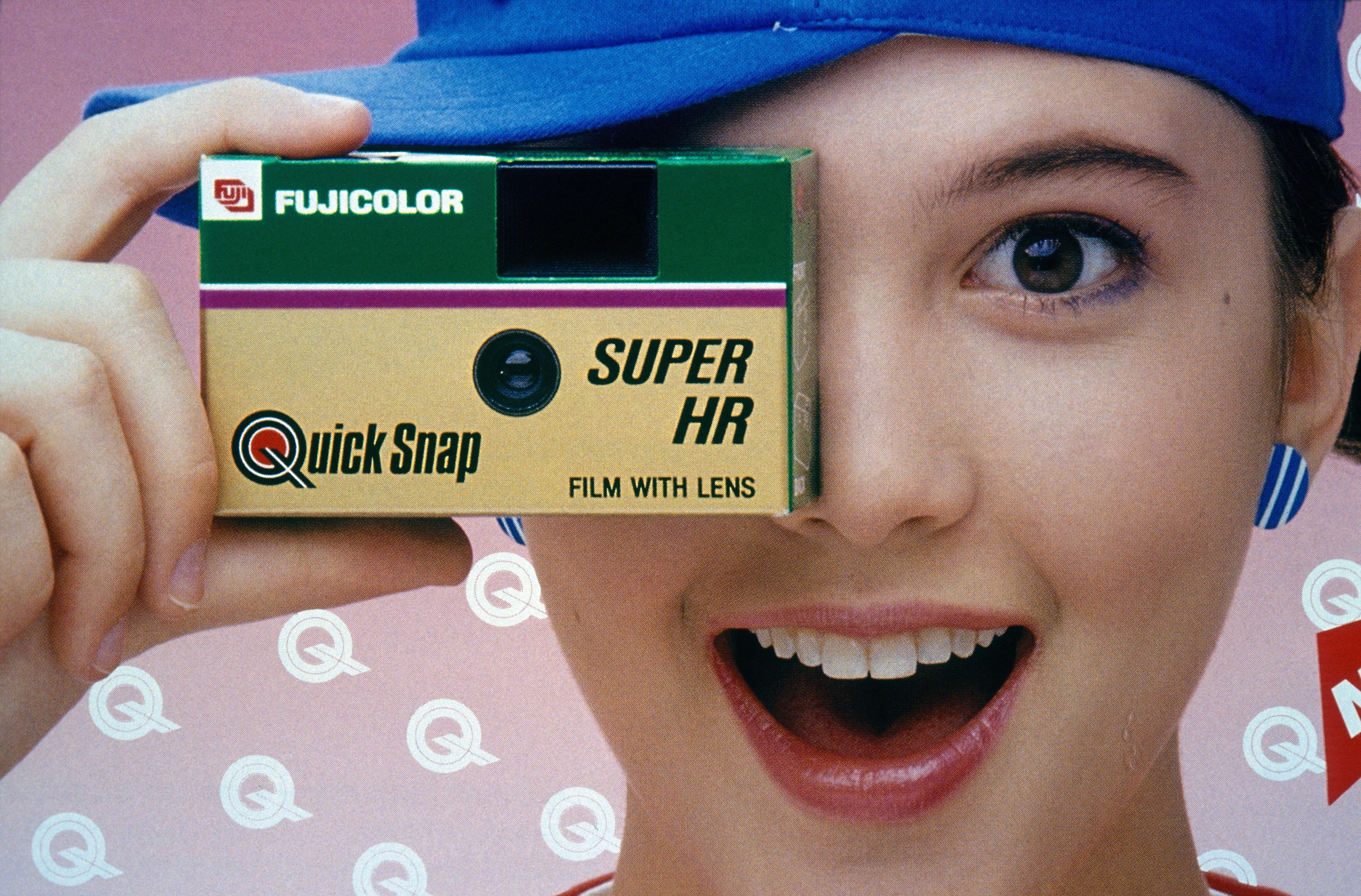 Person smiling, holding a Fujicolor QuickSnap camera to one eye against a patterned backdrop