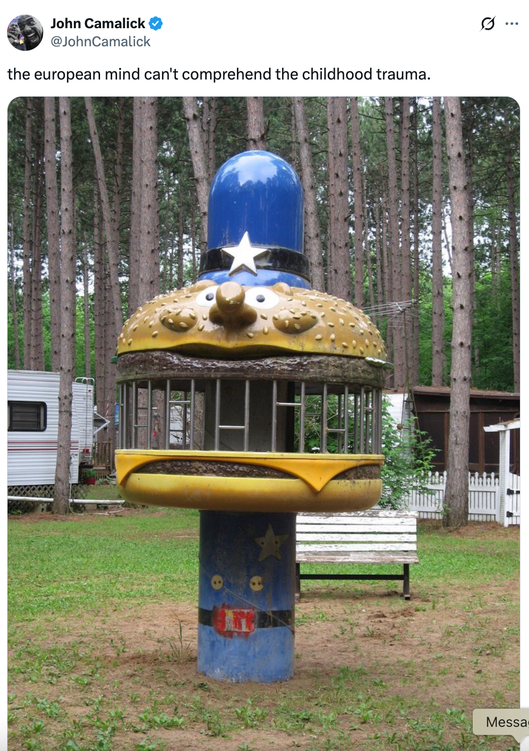 Large whimsical playground structure resembling a burger with a face, set in a wooded area with trailers nearby