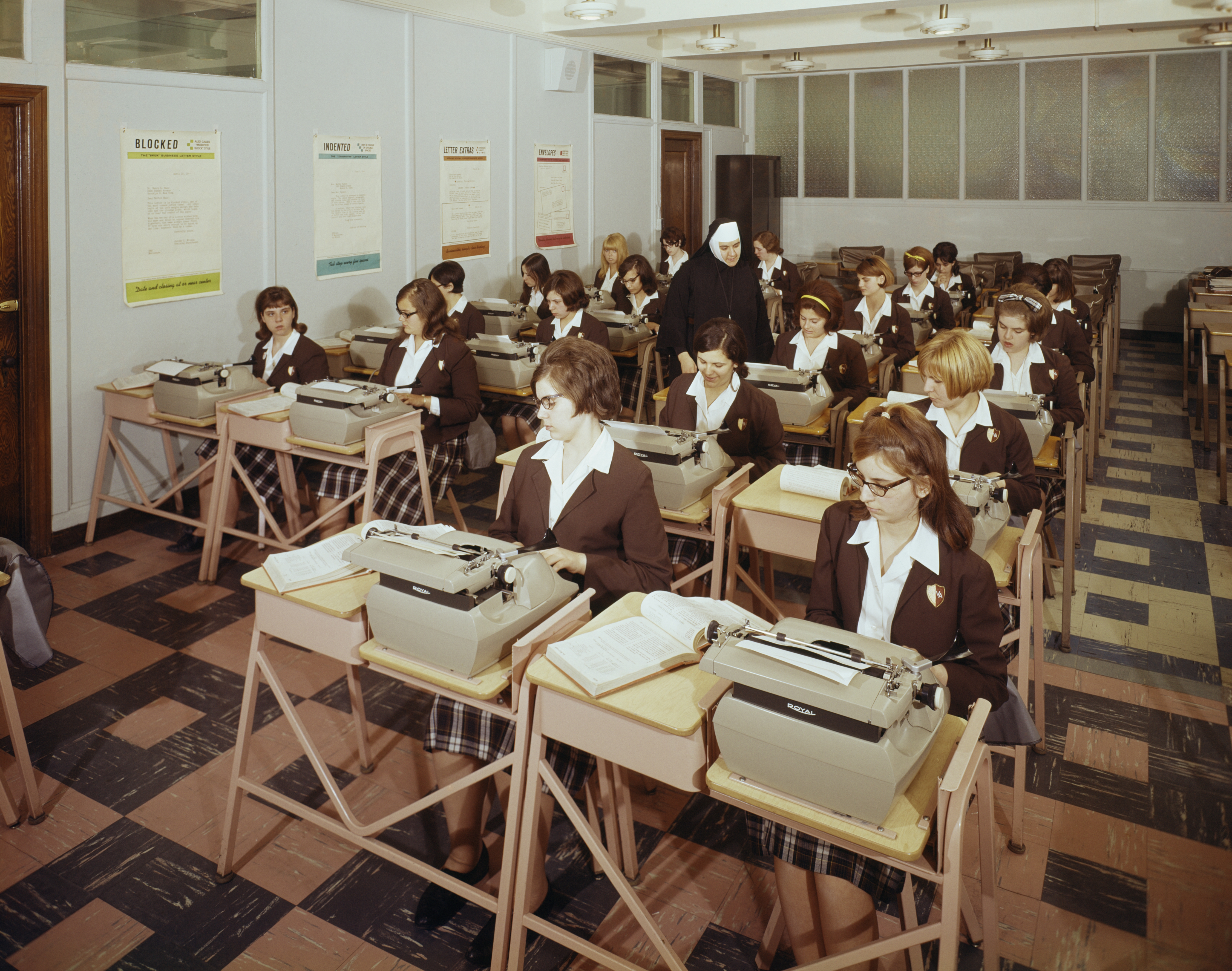 Students in uniforms sit at desks with typewriters in a classroom, guided by an instructor