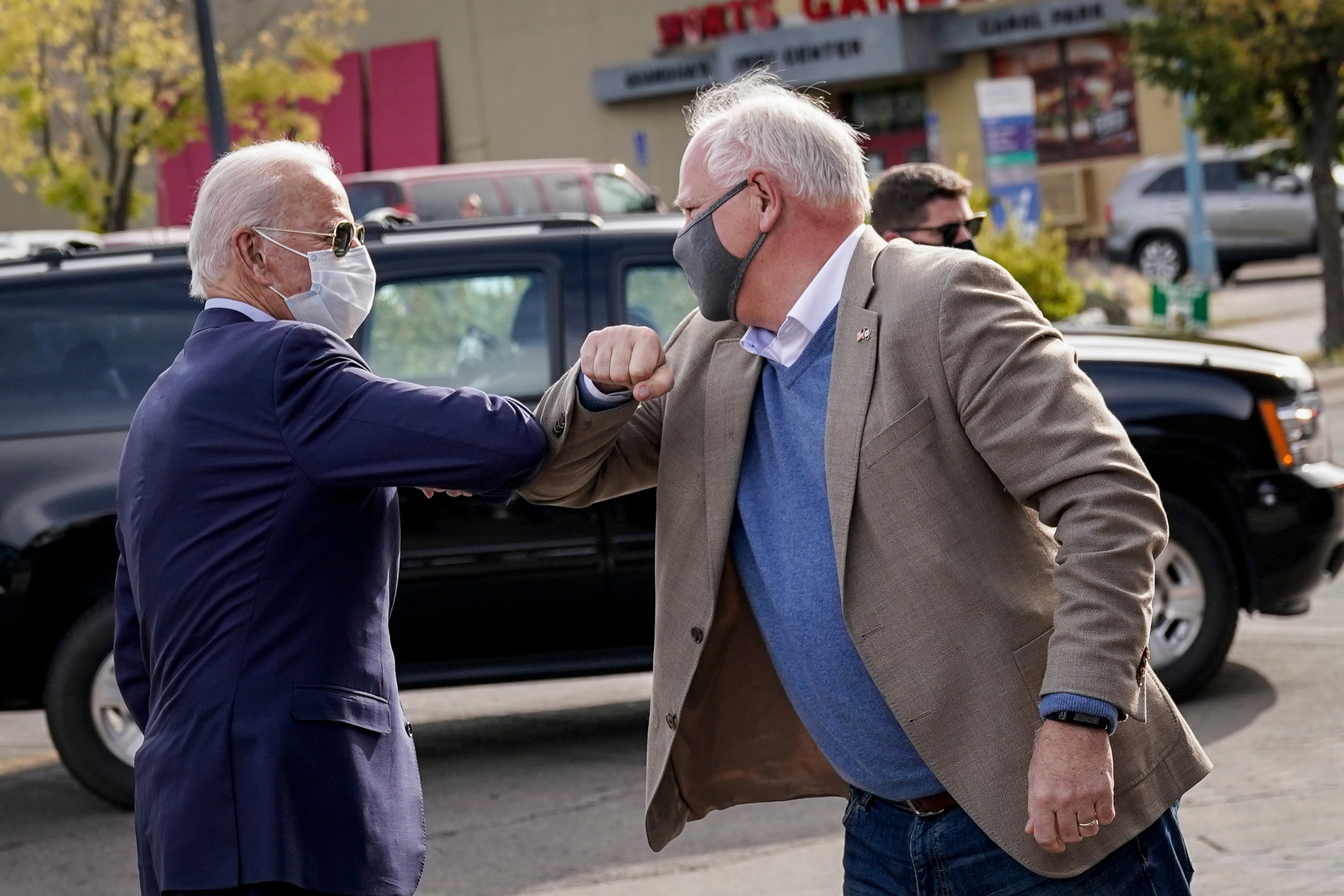 Two masked men greet each other with an elbow bump outdoors, near parked cars. Both are wearing casual jackets