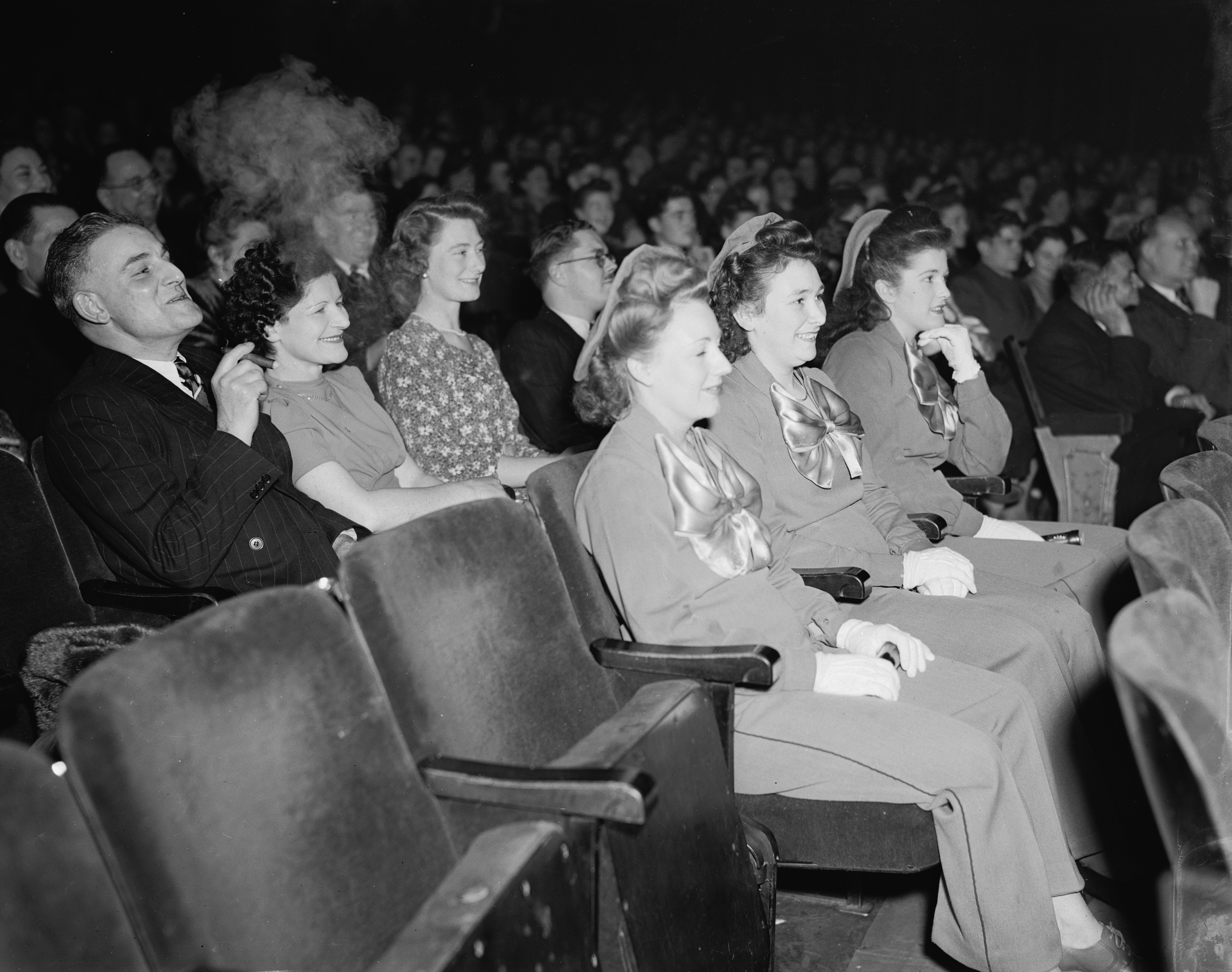 Audience members in a theater react with surprise and amusement while watching a performance, dressed in mid-20th-century attire
