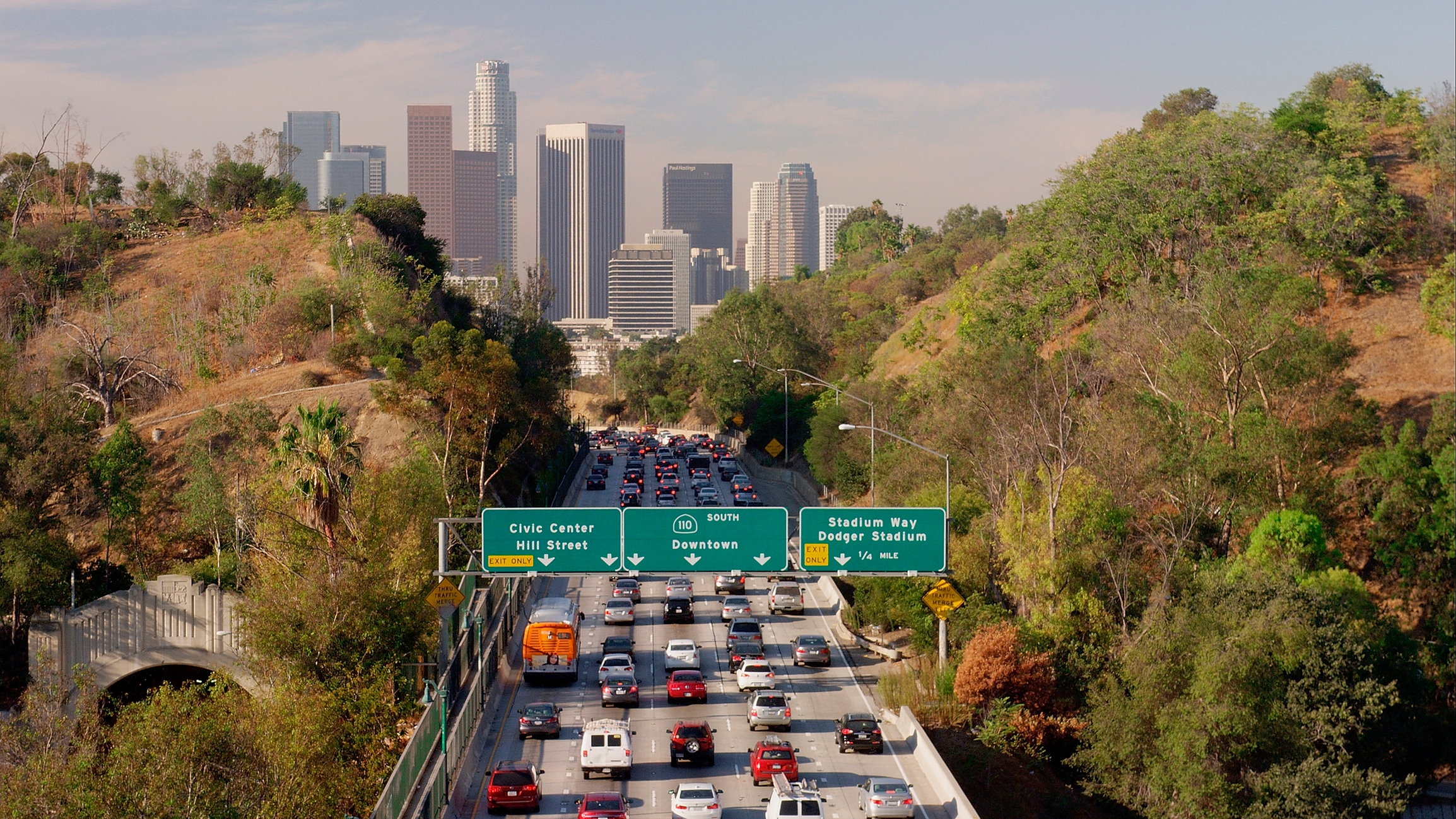 Heavy traffic on a highway with downtown Los Angeles skyscrapers in the background, surrounded by trees and hills