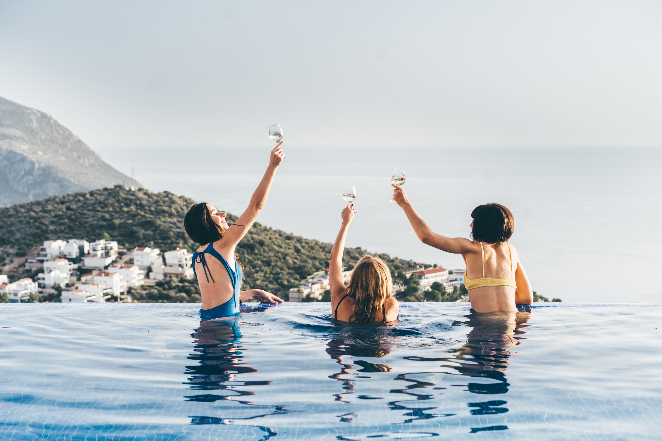 Three people in swimwear raise glasses while standing in an infinity pool overlooking a coastal view