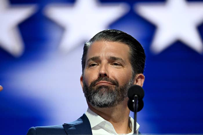 A person with slicked-back hair and a beard speaks at a podium, with large star shapes visible in the background
