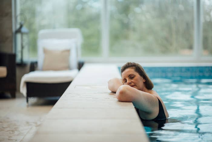 A woman relaxes and rests her head on the edge of an indoor pool, eyes closed, enjoying a peaceful moment