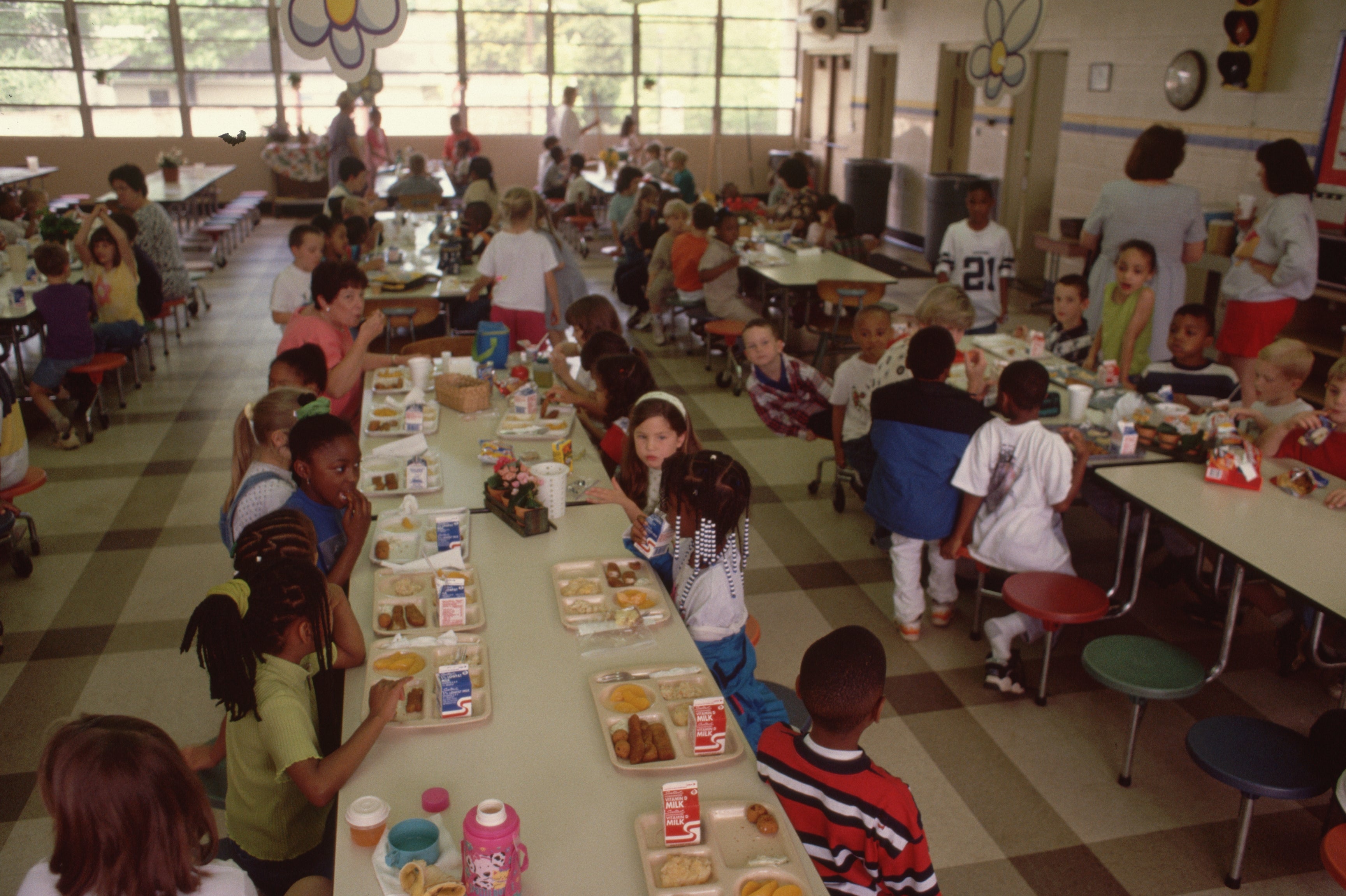 Students sitting at long cafeteria tables eating lunch, with a diverse group of children enjoying their meals and chatting