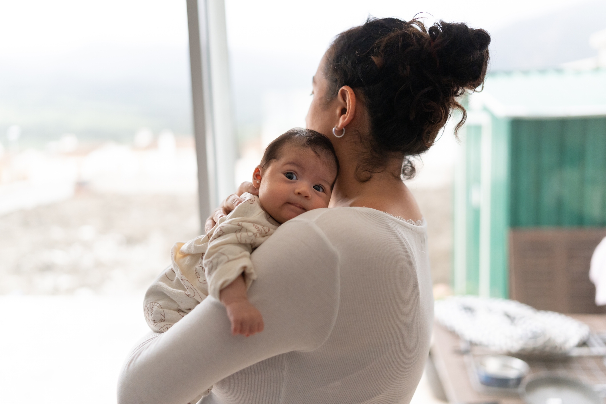 A woman stands by a window holding a baby on her shoulder, looking out. The baby is gazing towards the camera, wearing cozy clothing