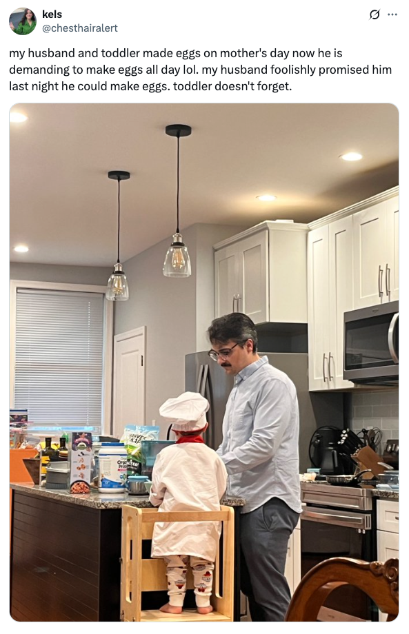 A man and a toddler wearing a chef's hat are in a kitchen, preparing food together