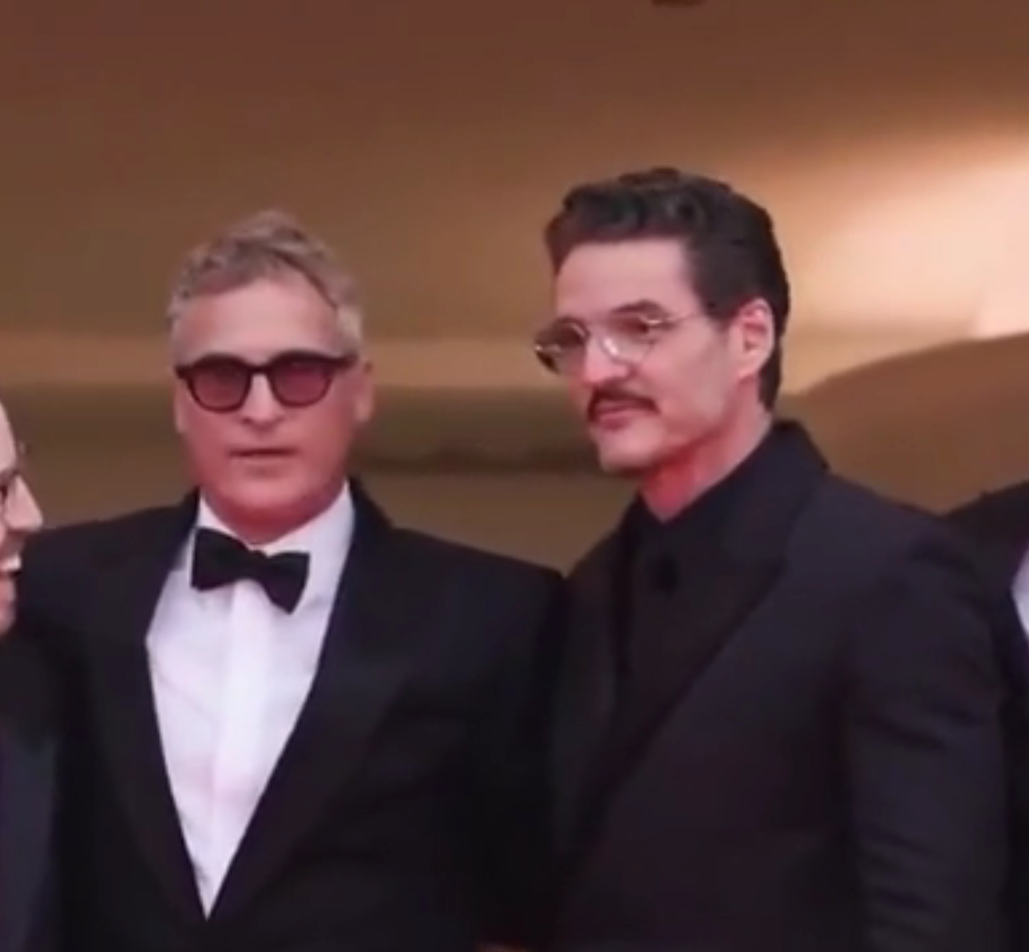 Two individuals on a red carpet, both in dark suits. One wears sunglasses. They pose for photos at an event