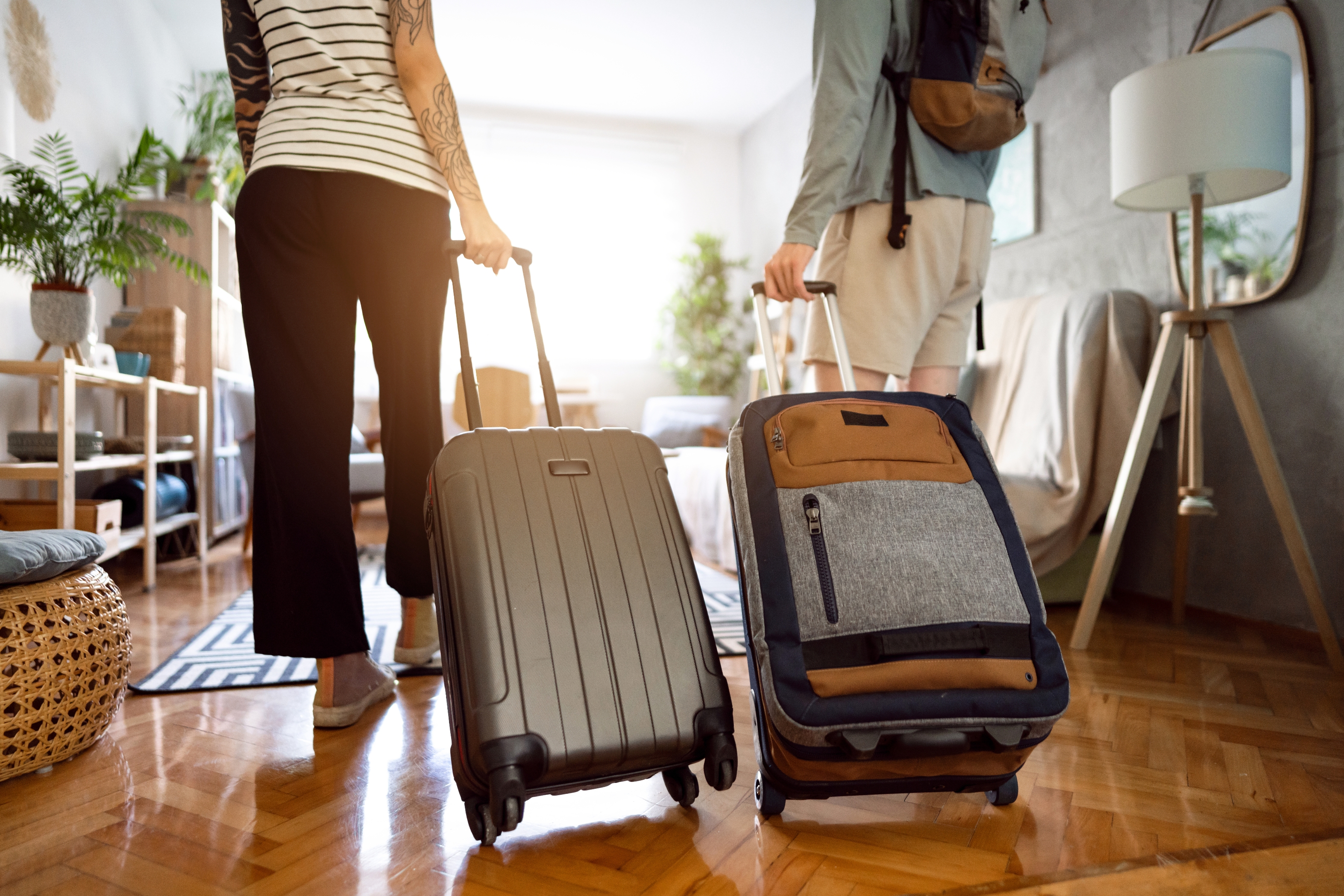 Two people with tattoos are pulling suitcases through a stylish, modern living room with wooden flooring and plants