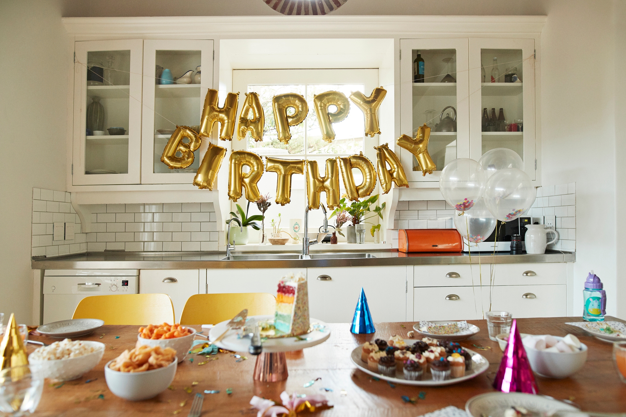 Kjøkken med & quot; Happy Birthday & quot; Ballonger, festhatter, snacks, cupcakes og en kake på et bord, som antyder en bursdagsfeiringsoppsett