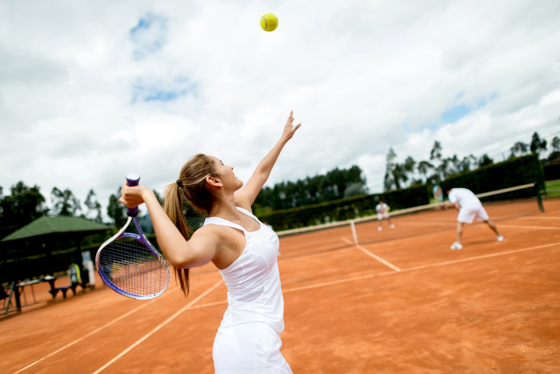 Person i tennisantrekk som serverer en ball på en utendørs leirbane, med en dobbeltpartner klar i bakgrunnen