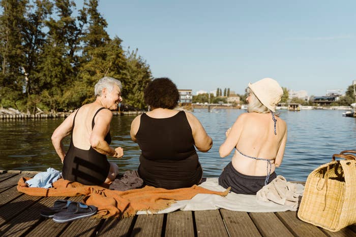 Three women sit on a dock by a lake, talking and laughing under clear skies