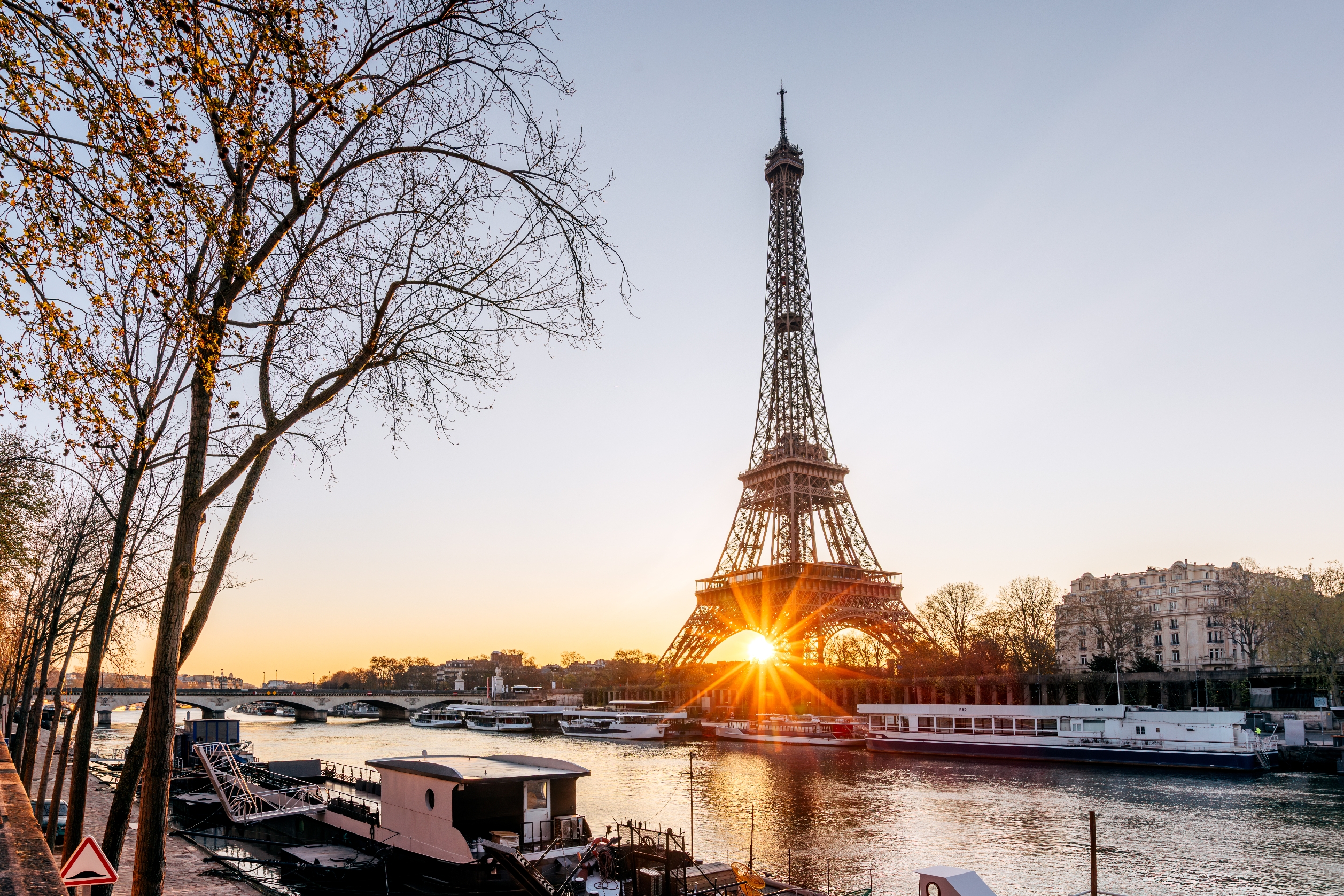 Eiffel Tower with sun setting below, casting reflections on the Seine River; a scenic Parisian view with trees along the riverbank
