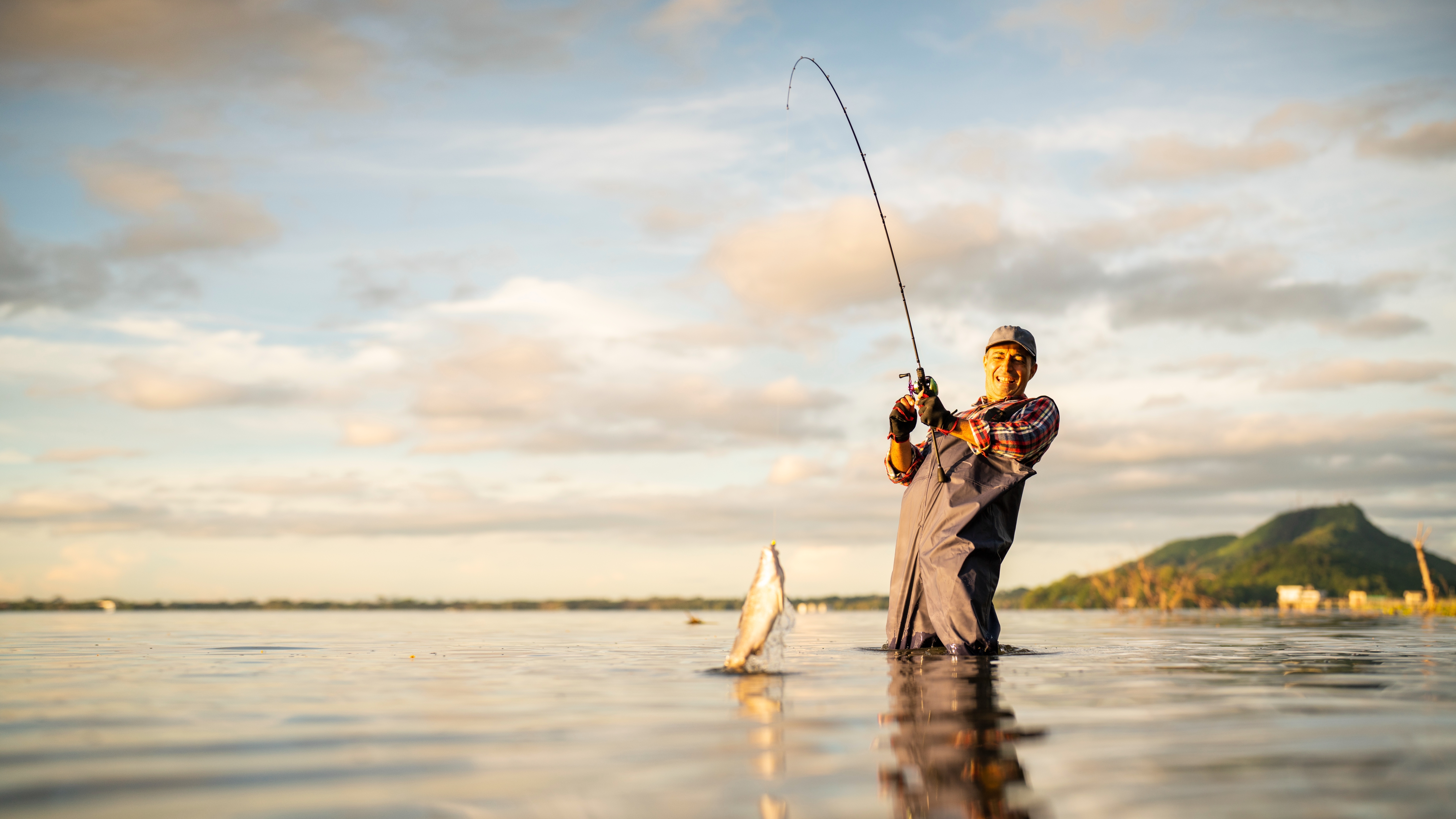 Person wearing a cap and overalls stands in water while catching a fish, appearing joyful and focused