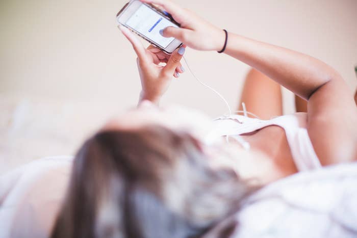 Person lying down, using a smartphone with headphones. Their face is not visible, and they appear relaxed, focusing on the device