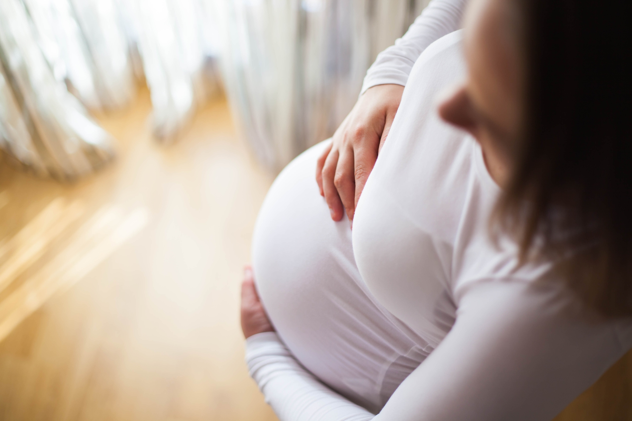 Gravid person som har en langermet topp forsiktig vugger magen og står i et mykt opplyst rom