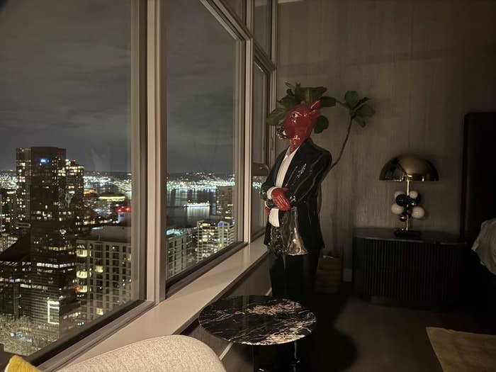 A stylishly dressed person with a bull's head mask stands by a large window, overlooking a cityscape at night from a modern apartment