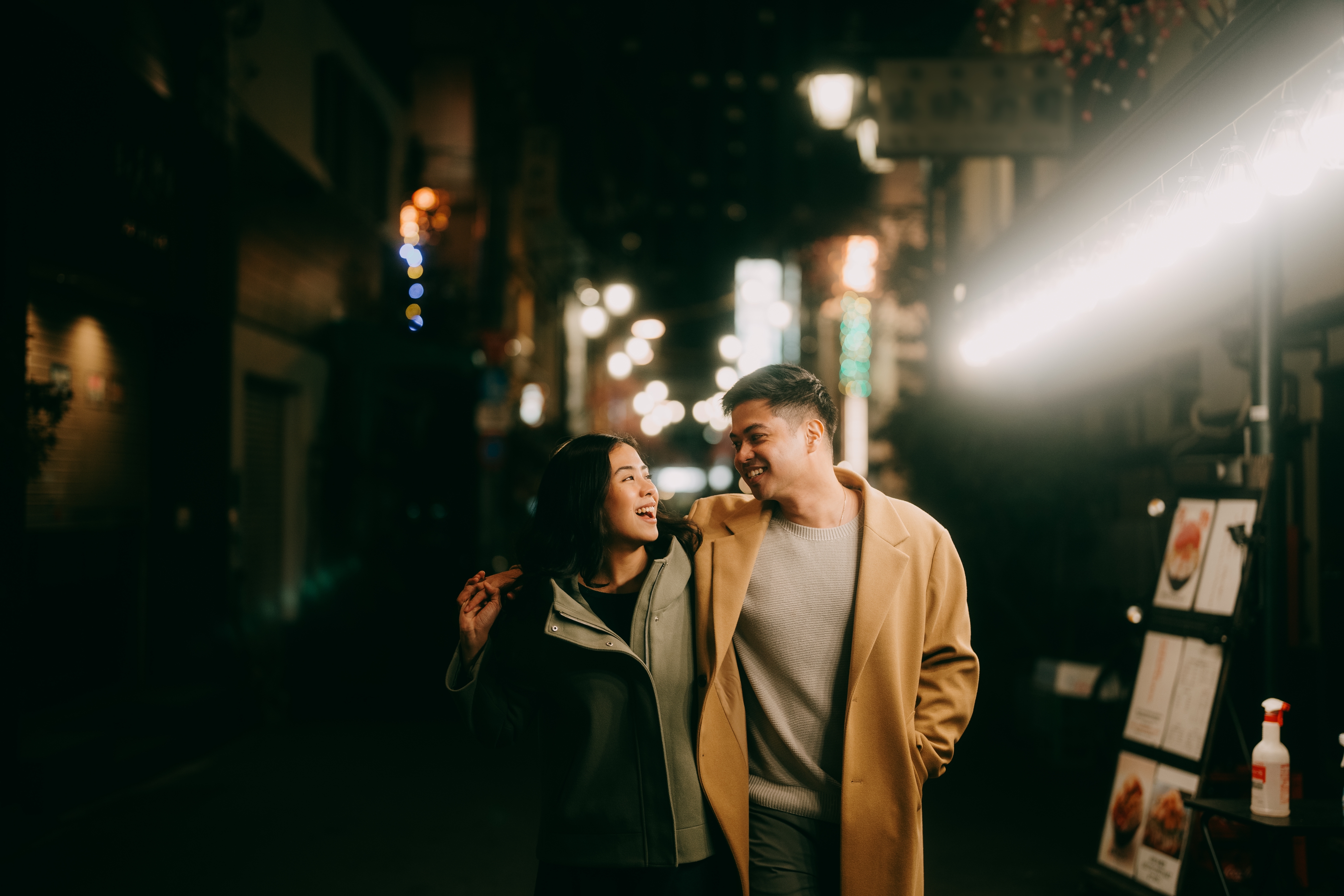 A couple in stylish coats smiles at each other, walking closely at night, illuminated by streetlights
