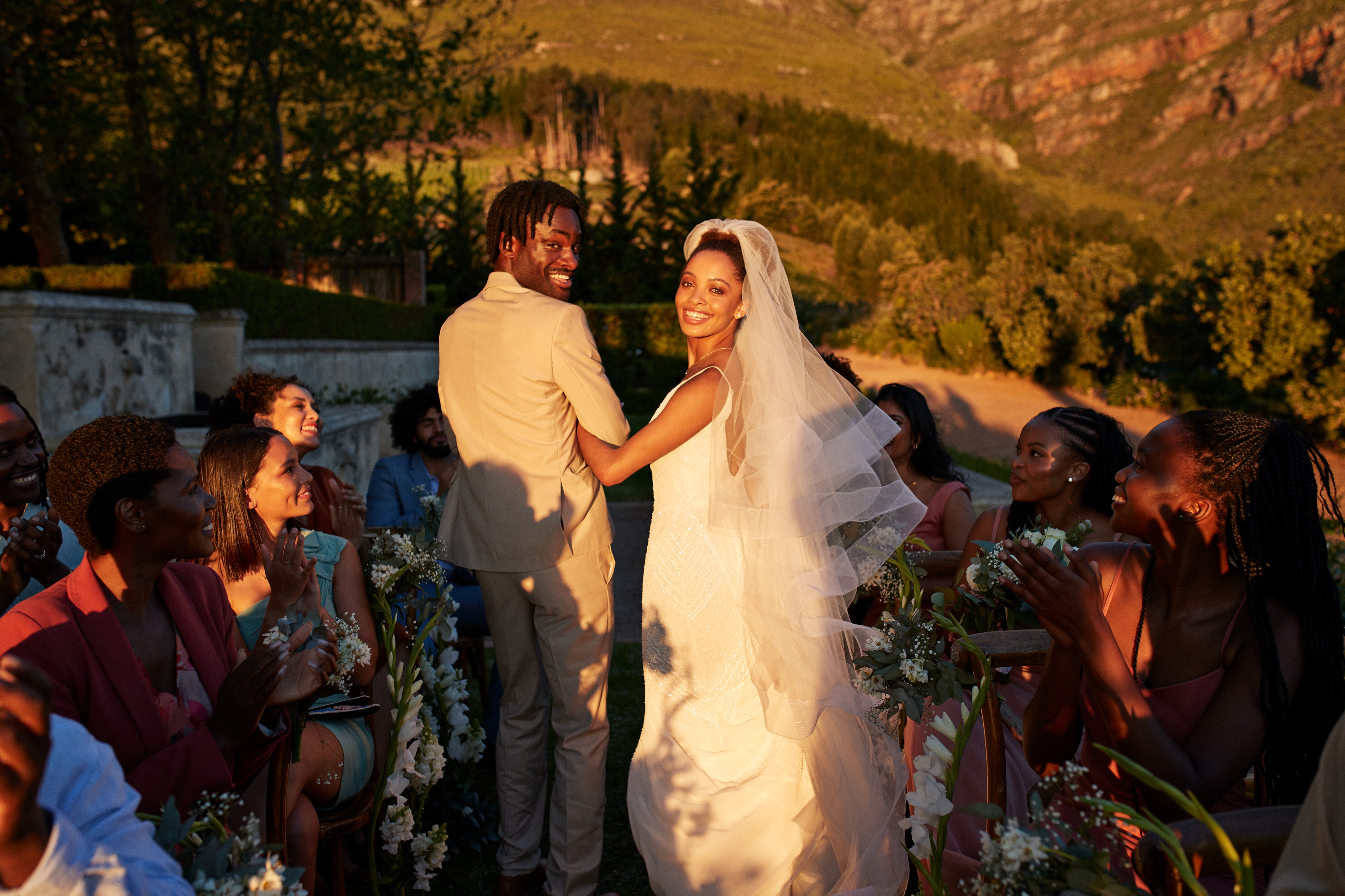 A joyful couple in wedding attire walks down an aisle outdoors, surrounded by smiling guests, capturing a moment of love and celebration