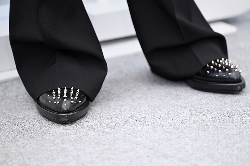 Person wearing wide black pants and shoes with sharp metal spikes on the toes, standing on a light-colored surface