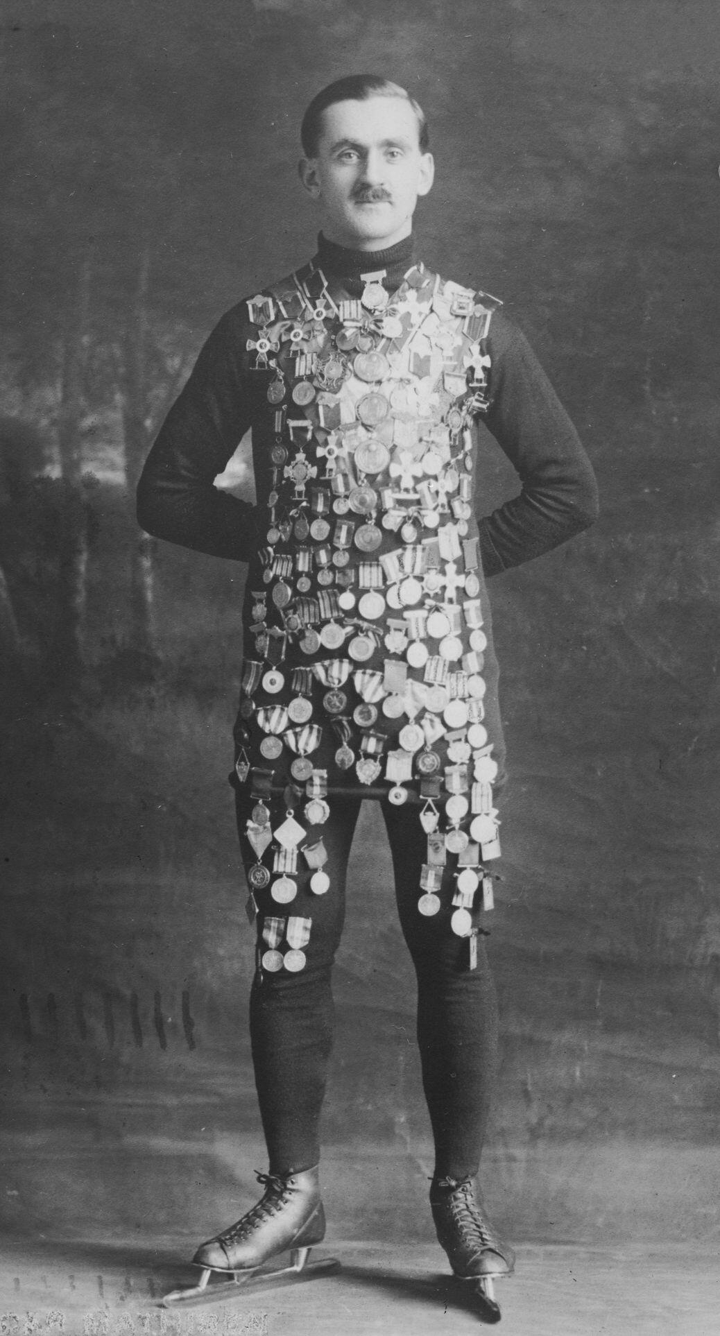 A man stands wearing numerous medals covering his torso on top of a dark outfit, with his hands behind his back and expressionless face