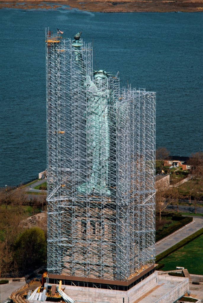 The Statue of Liberty surrounded by scaffolding for refurbishment, with a view of the water and nearby land