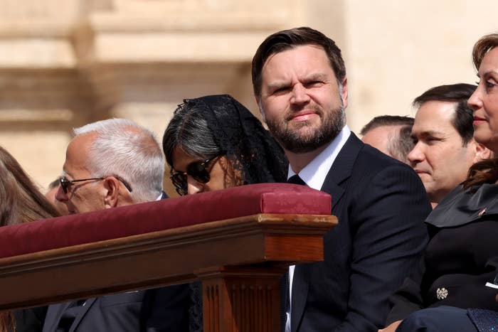 A man in a suit sits with a group at an outdoor event, looking toward the distance. Other attendees are partially visible